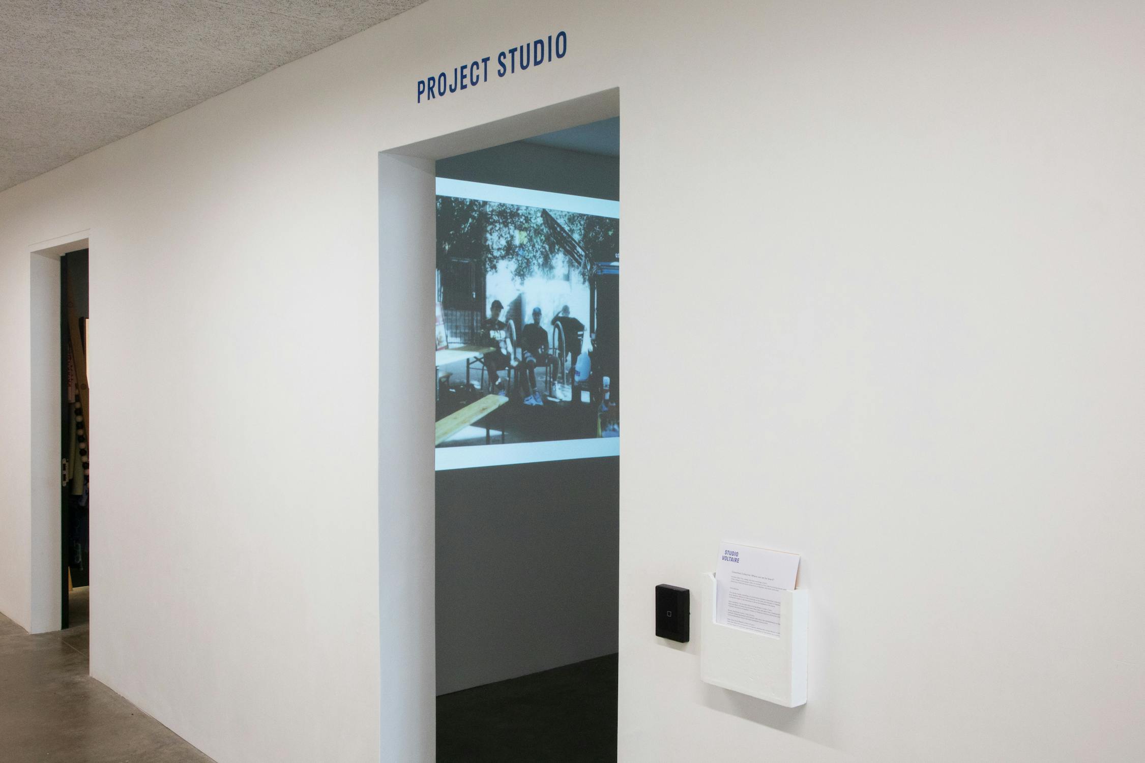 A white corridor with a doorway labelled “PROJECT STUDIO.” Inside the doorway, a projection of a black-and-white image is displayed on the interior wall, showing people gathered outdoors. The wall outside has a white device and a black panel mounted on it.
