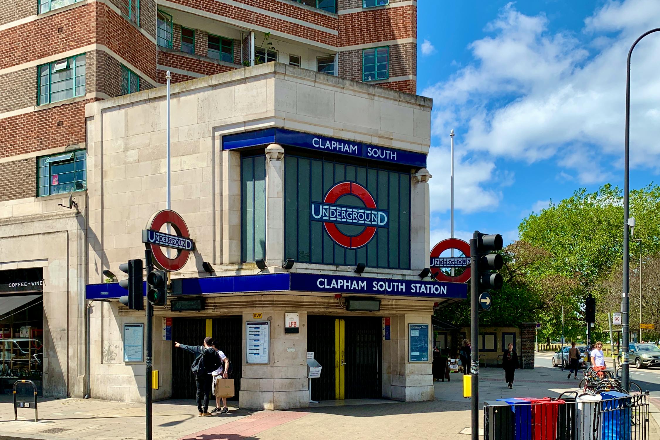 The image shows the entrance to Clapham South Station, a London Underground station. The building has a stone façade with two round red and blue Underground signs, and there is a person with a suitcase near the entrance on a sunny day. Trees are visible to the right.