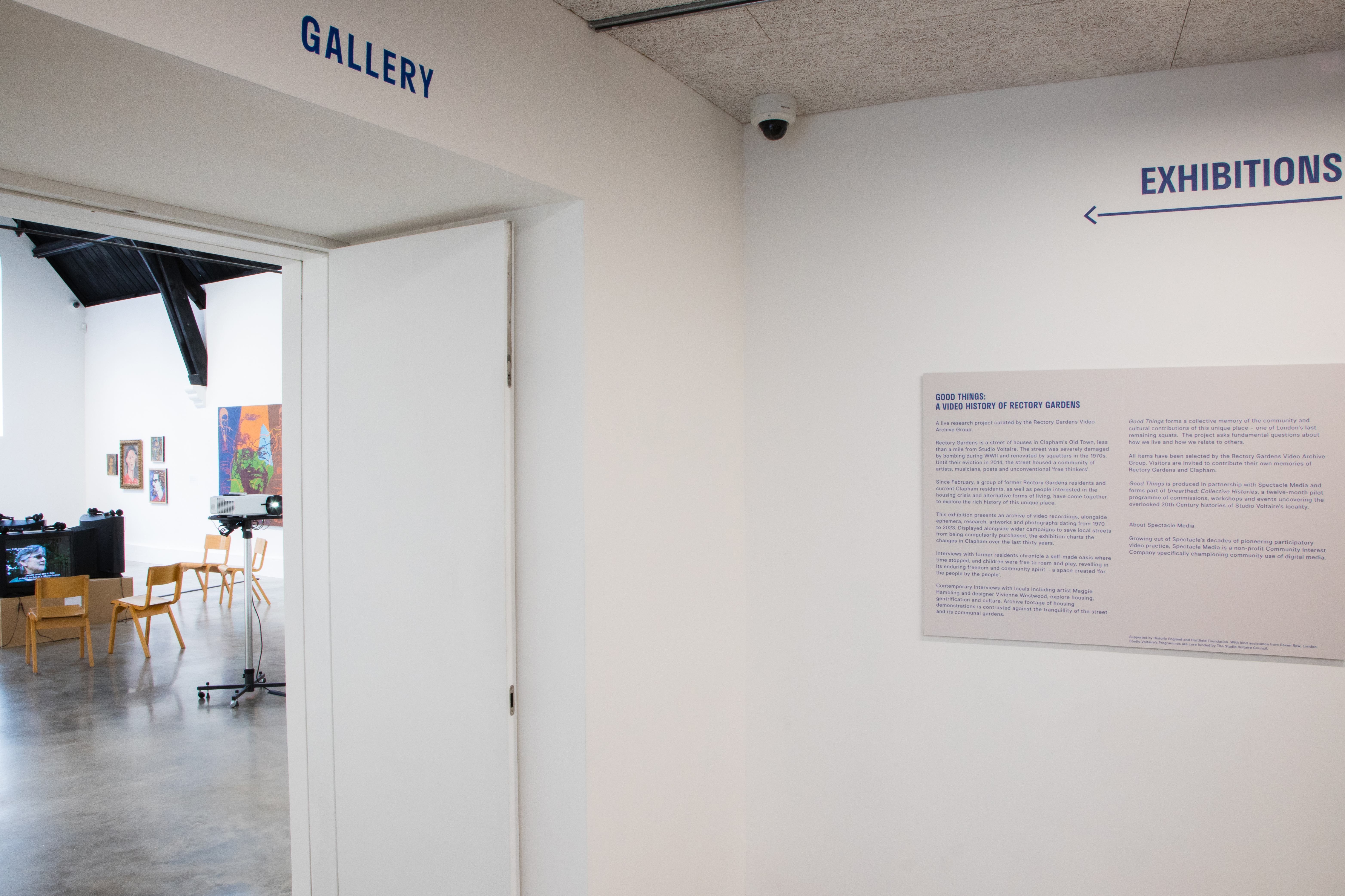 A white-walled gallery entrance with “GALLERY” and “EXHIBITIONS” signs in blue above doorways. A wall-mounted text panel is visible. Inside the gallery, there are paintings, tables, chairs, and equipment on a polished concrete floor.