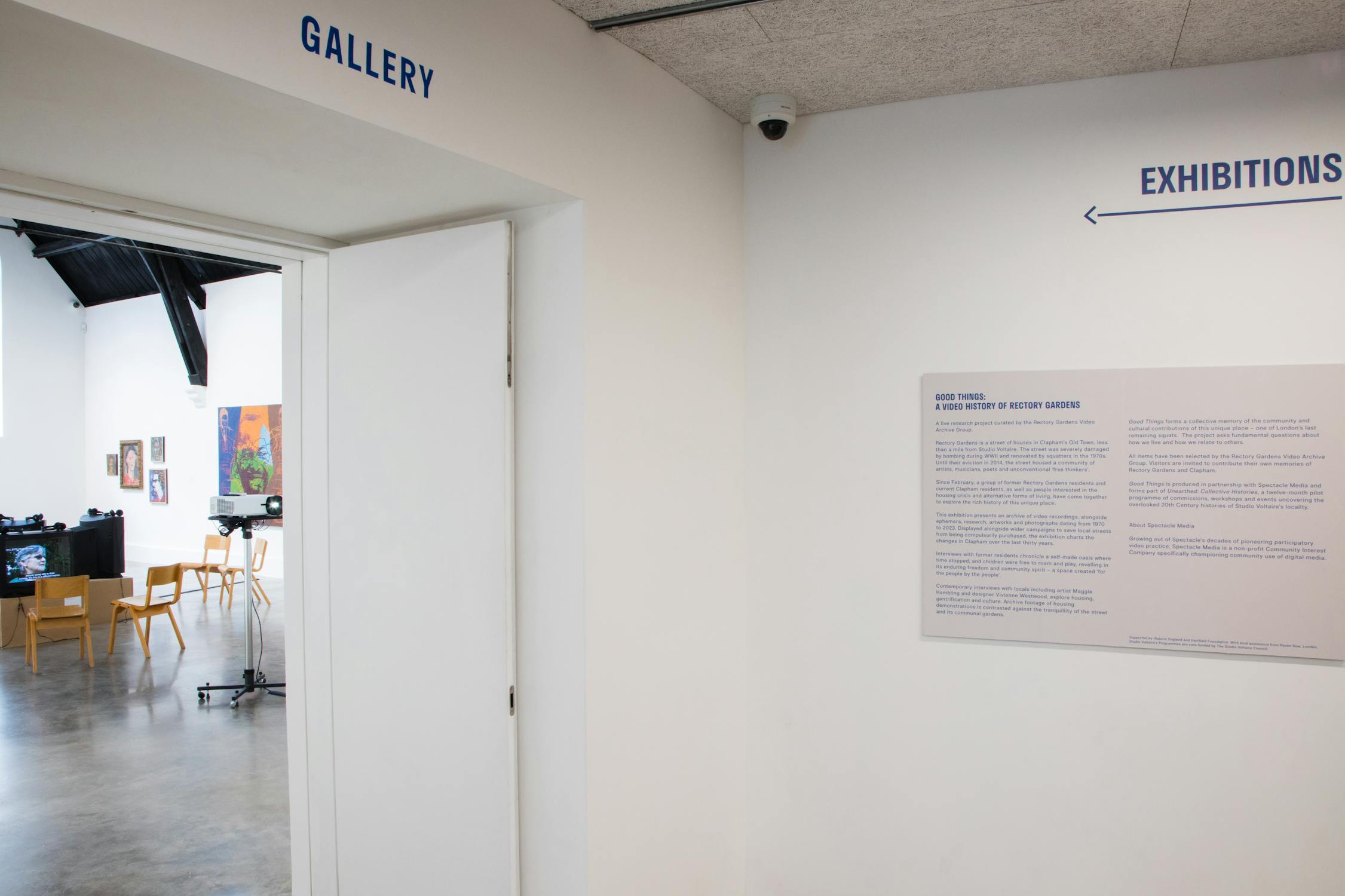 A white-walled gallery entrance with “GALLERY” and “EXHIBITIONS” signs in blue above doorways. A wall-mounted text panel is visible. Inside the gallery, there are paintings, tables, chairs, and equipment on a polished concrete floor.
