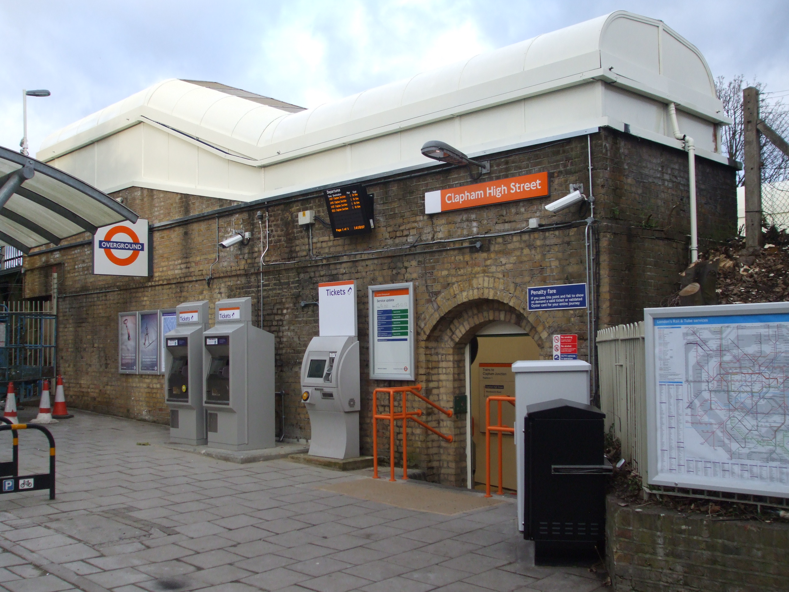Exterior view of a London Underground station with a brick façade. Signs read “Clapham High Street Station.” Ticket machines, an information board, and a map are visible outside. The entrance has an orange sign, and the pavement is empty.