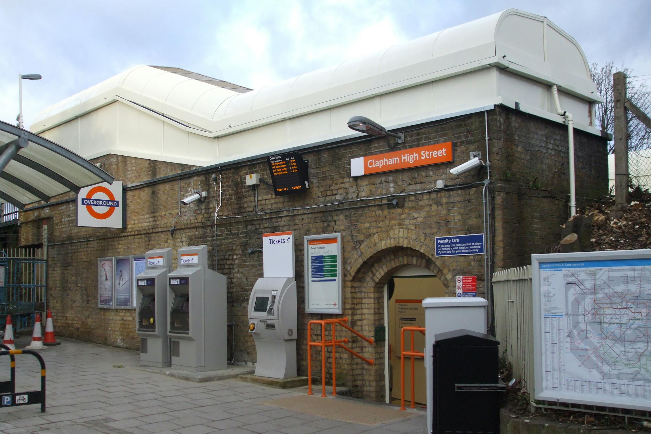 Exterior view of a London Underground station with a brick façade. Signs read “Clapham High Street Station.” Ticket machines, an information board, and a map are visible outside. The entrance has an orange sign, and the pavement is empty.