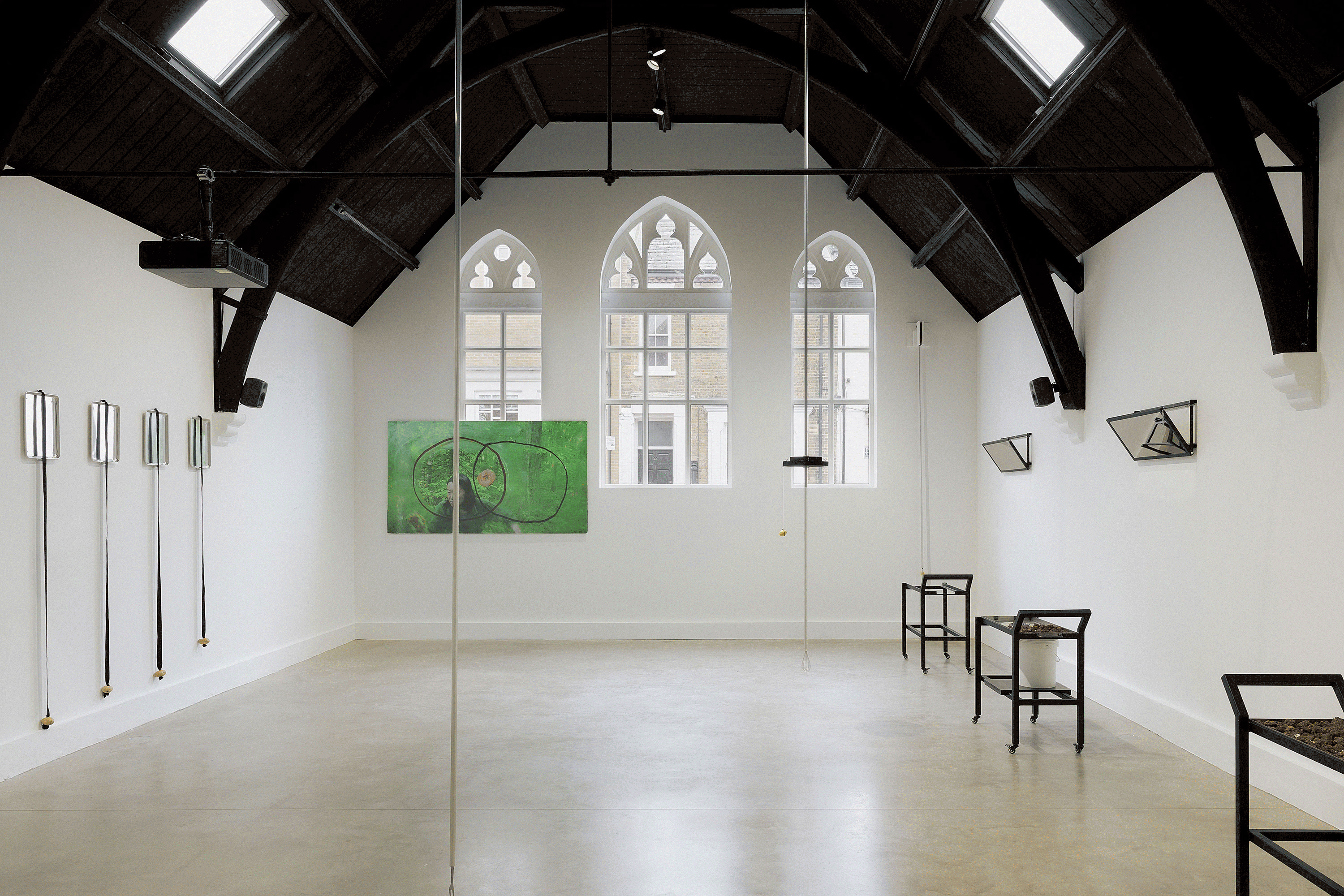 Two images – the first image shows a bright, minimalist gallery. Contemporary art, including a green abstract painting and metallic installations, is displayed along the walls. The room has polished concrete floors. The second image shows a dimly lit gallery room featuring two folding chairs facing a large projected portrait of a seated woman in a historic dress and a small diorama depicting a figure in a forest-like setting. 