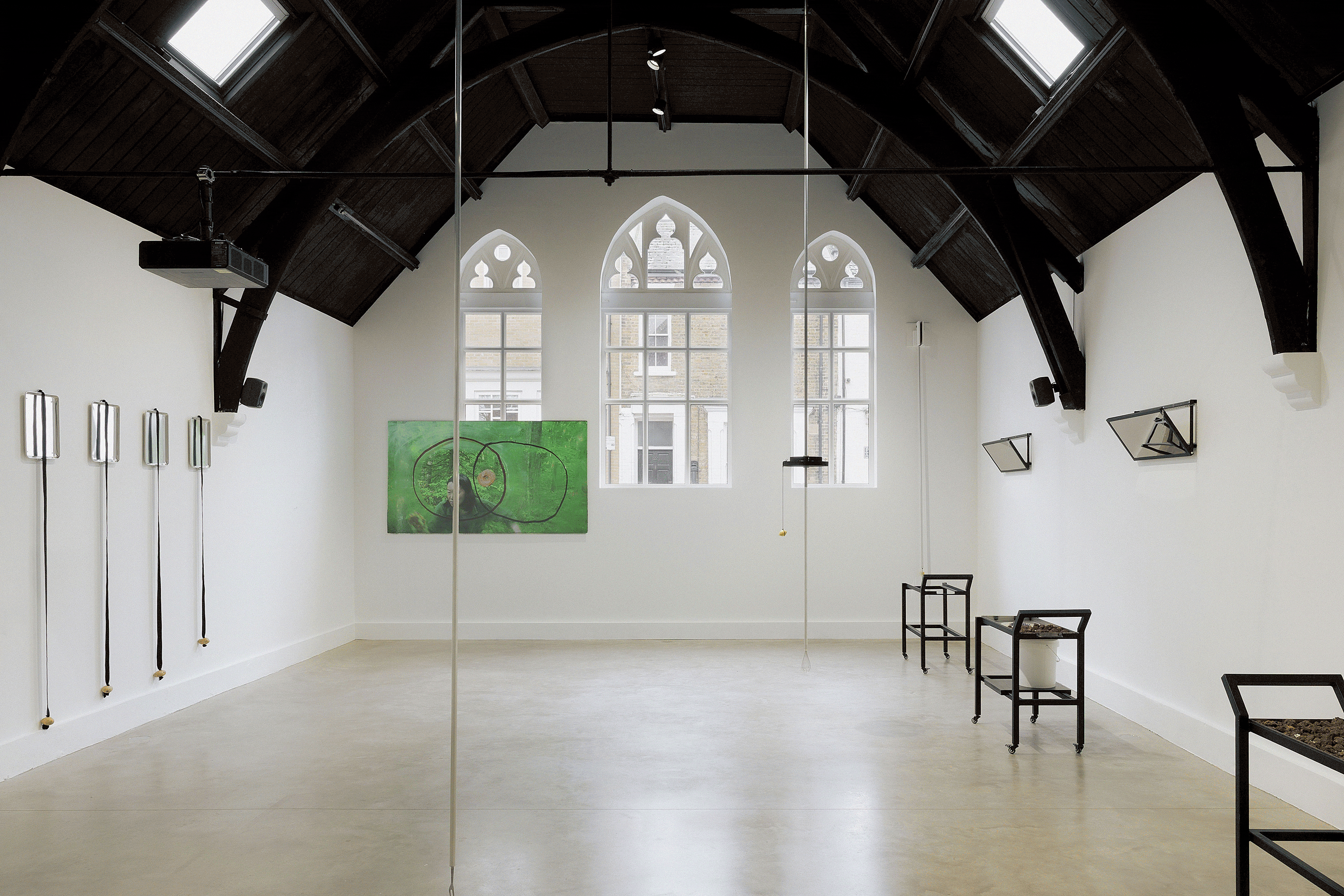 Two images – the first image shows a bright, minimalist gallery. Contemporary art, including a green abstract painting and metallic installations, is displayed along the walls. The room has polished concrete floors. The second image shows a dimly lit gallery room featuring two folding chairs facing a large projected portrait of a seated woman in a historic dress and a small diorama depicting a figure in a forest-like setting.
