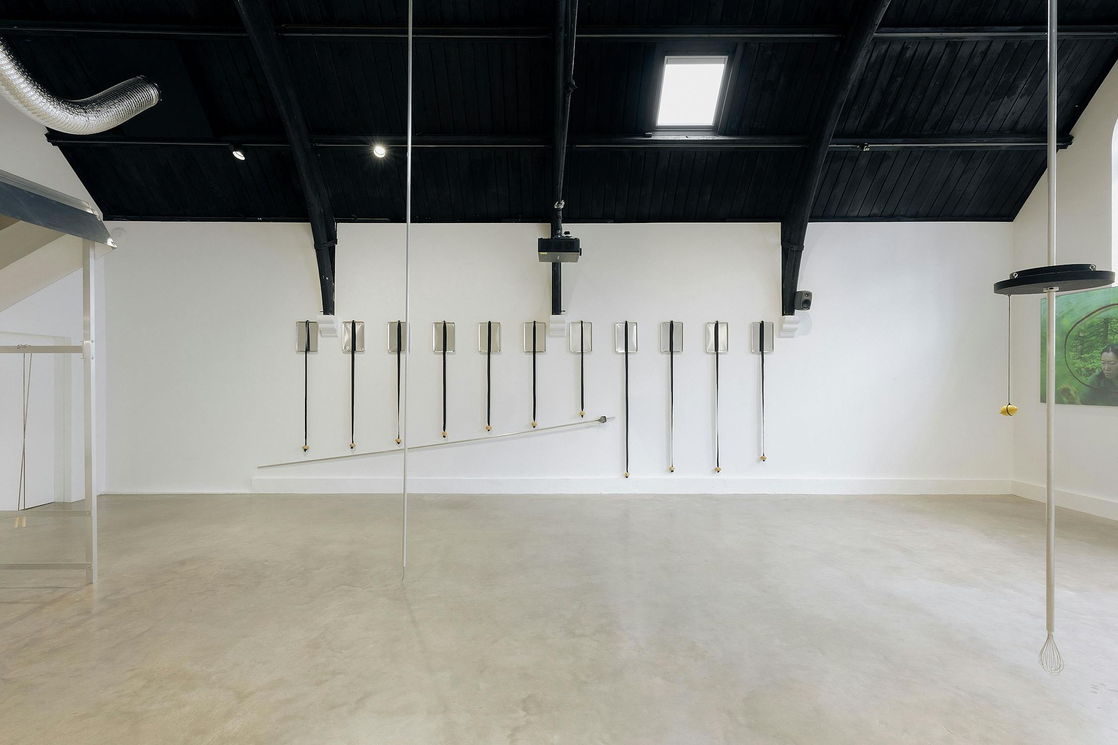 A minimalist gallery with a polished concrete floor, white walls, and a black vaulted ceiling. On the wall, potatoes on rods are arranged in a line, descending diagonally. Overhead pipes and a projector are also visible.