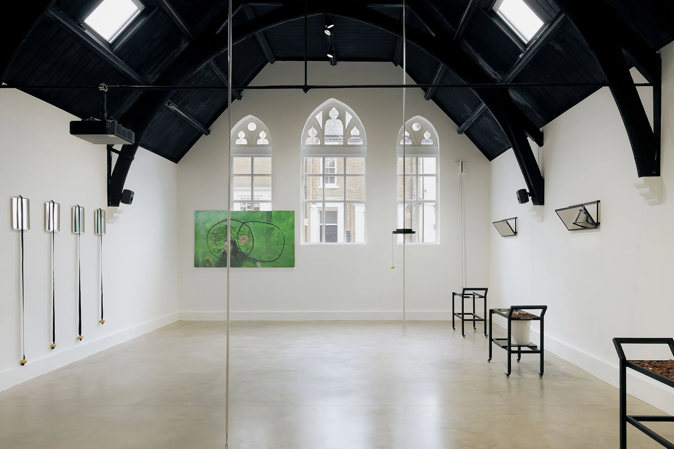 A modern art gallery with high, dark vaulted ceilings, arched windows, and white walls. Minimalist sculptures are mounted on the walls, including long rods and mirrors. A green abstract painting hangs between the windows. Sparse black tables stand along the sides.