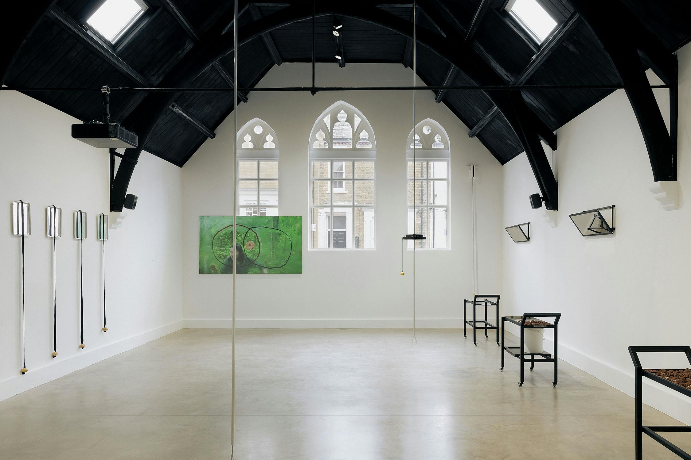 A contemporary art gallery with high, dark vaulted ceilings and arched windows. Minimalist sculptures and framed artworks are displayed on white walls and shelves, with natural light streaming in. The space appears clean, open, and modern.