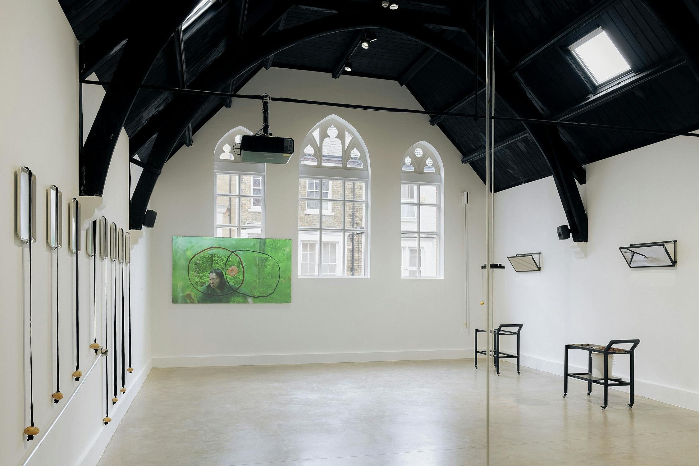A modern art gallery with high arched ceilings, tall windows, and white walls. Minimalist sculptures and objects are displayed on the walls and on black tables, with a green abstract painting between two windows at the far end. The space is bright and airy.