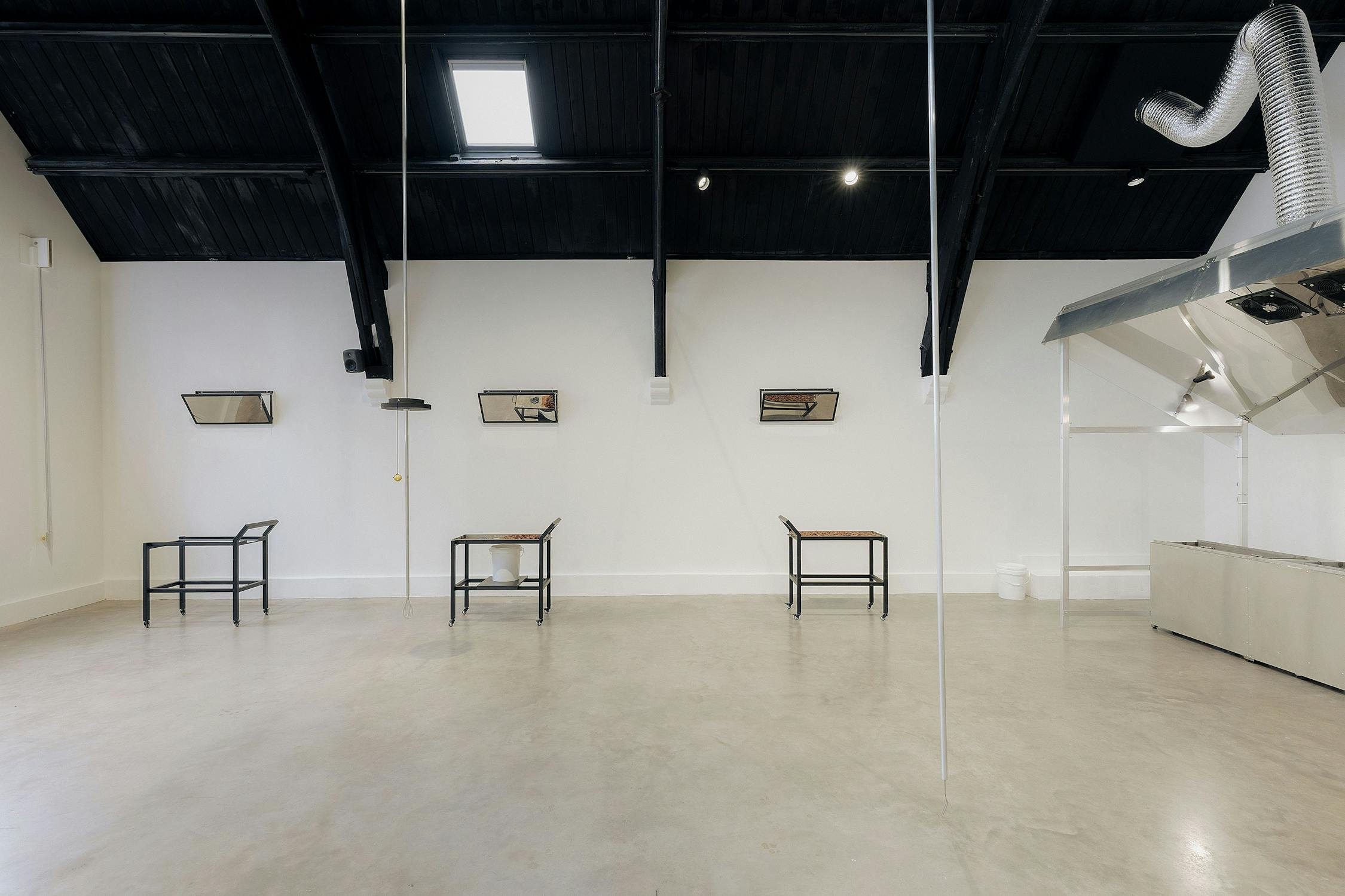 A minimalist gallery with high, black-beamed ceilings, white walls, and arched windows. Three black metal tables with patterned surfaces are spaced along the wall beneath three angled, glass-fronted display cases. The space is bright and uncluttered.