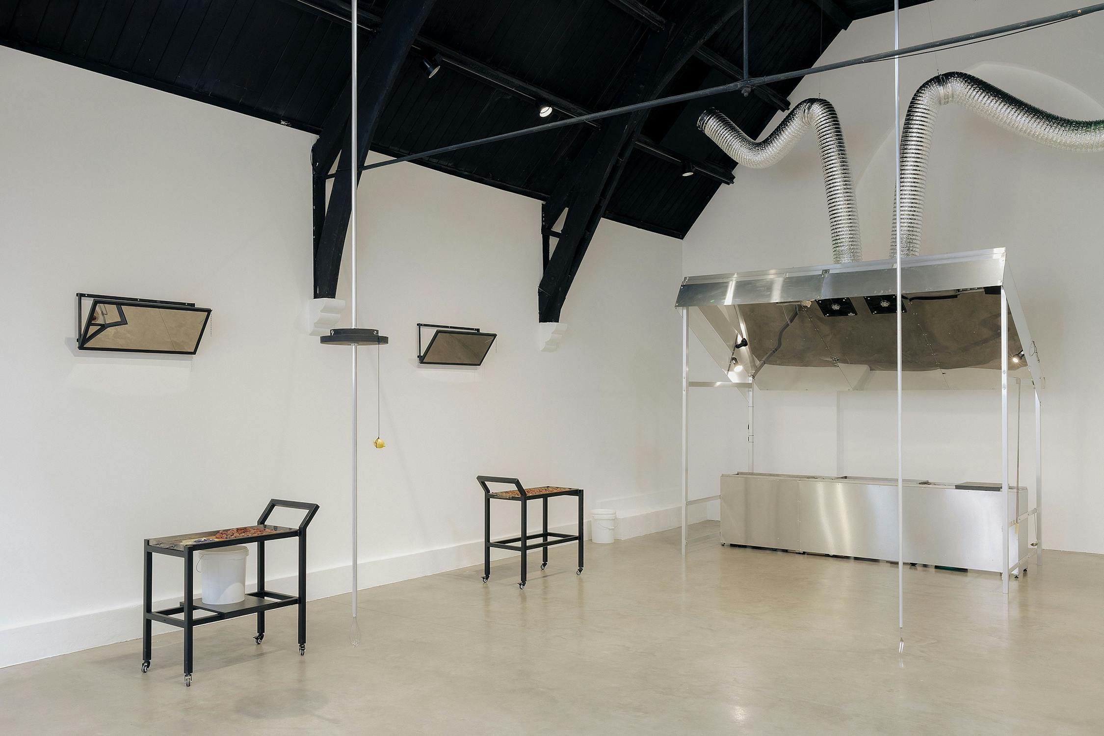 A minimalist room with a high black ceiling, white walls, and a polished concrete floor. Two simple tables with chairs face the wall, each with a rectangular mirror above. On the right, there is a metal structure with ventilation pipes. The space is open and modern.