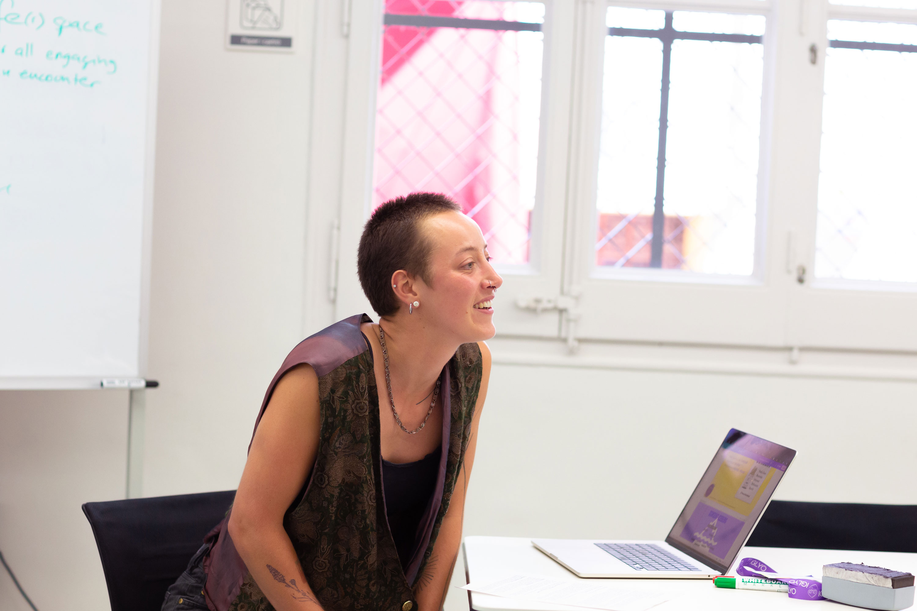Ron Eis, with short hair and sleeveless clothing smiles while leaning forward at a desk with a laptop, notebook, and markers. Behind them are large windows with bars and a whiteboard partially visible on the left.