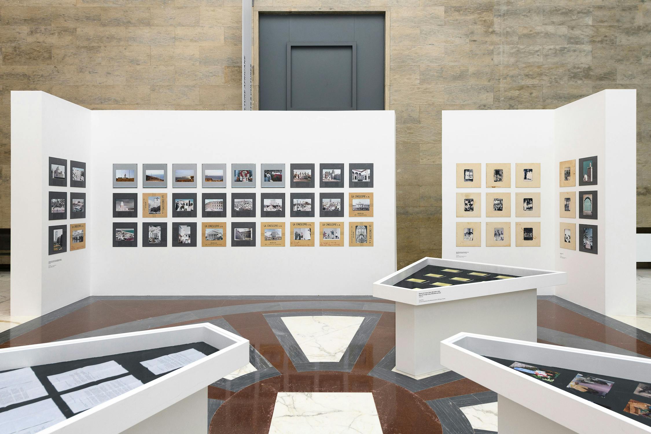 A modern gallery displays Georges Senga's art on white panels: photographs and small illustrations are arranged in rows. Two display tables with documents and objects are placed in the foreground on a glossy marble floor. The background features a tall beige wall.