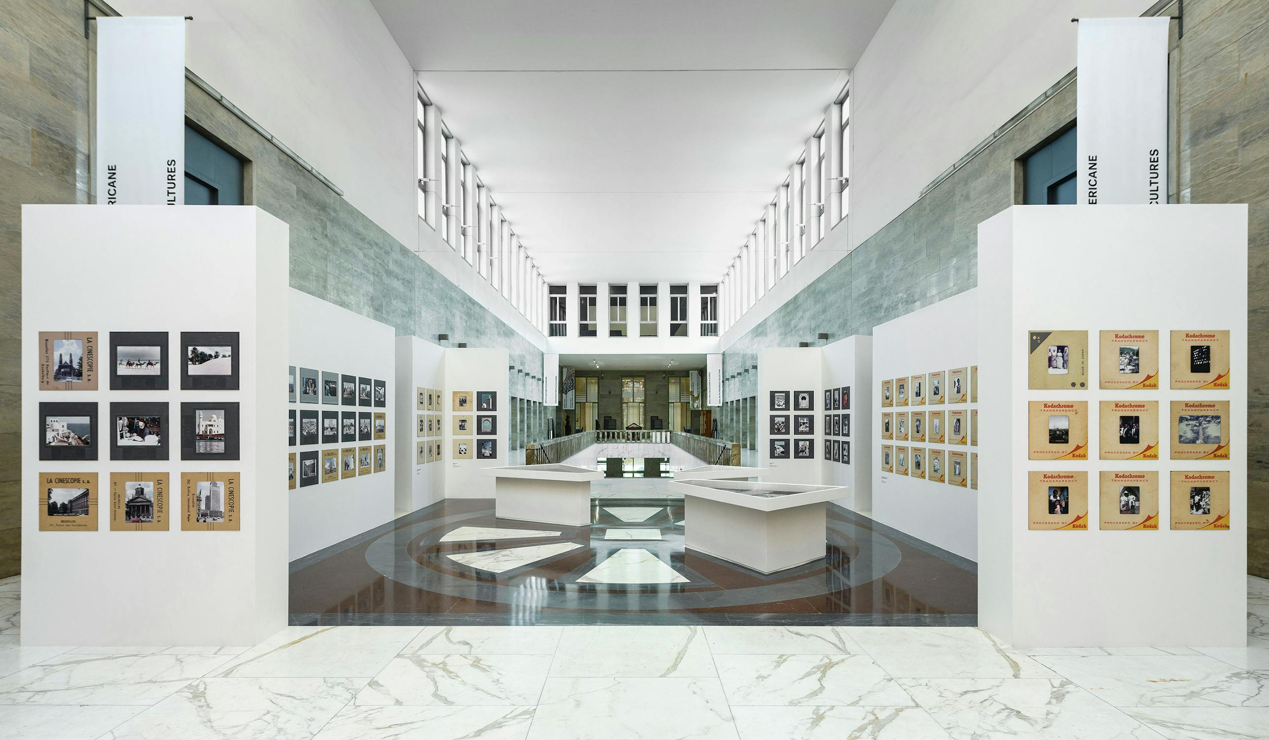 A modern gallery space with marble floors and high ceilings displays multiple white panels featuring framed photographs. In the centre, angular display tables hold more items. The space is bright, with large windows at the far end letting in natural light.