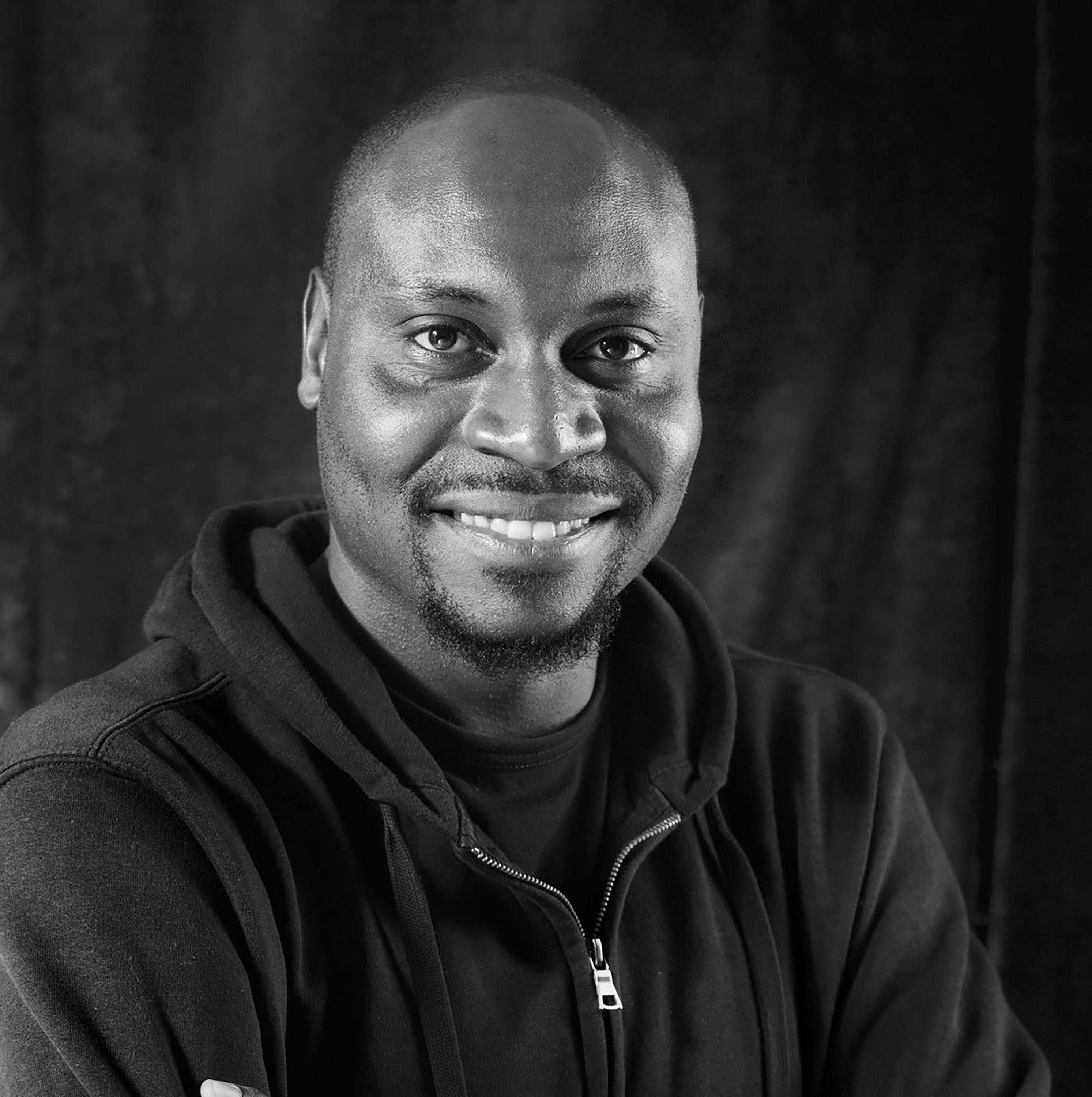 Portrait of Georges Senga. A black man with a shaven head and a short goatee smiles while looking at the camera. He wears a dark zip-up hoodie over a T-shirt. The photo is in black and white.