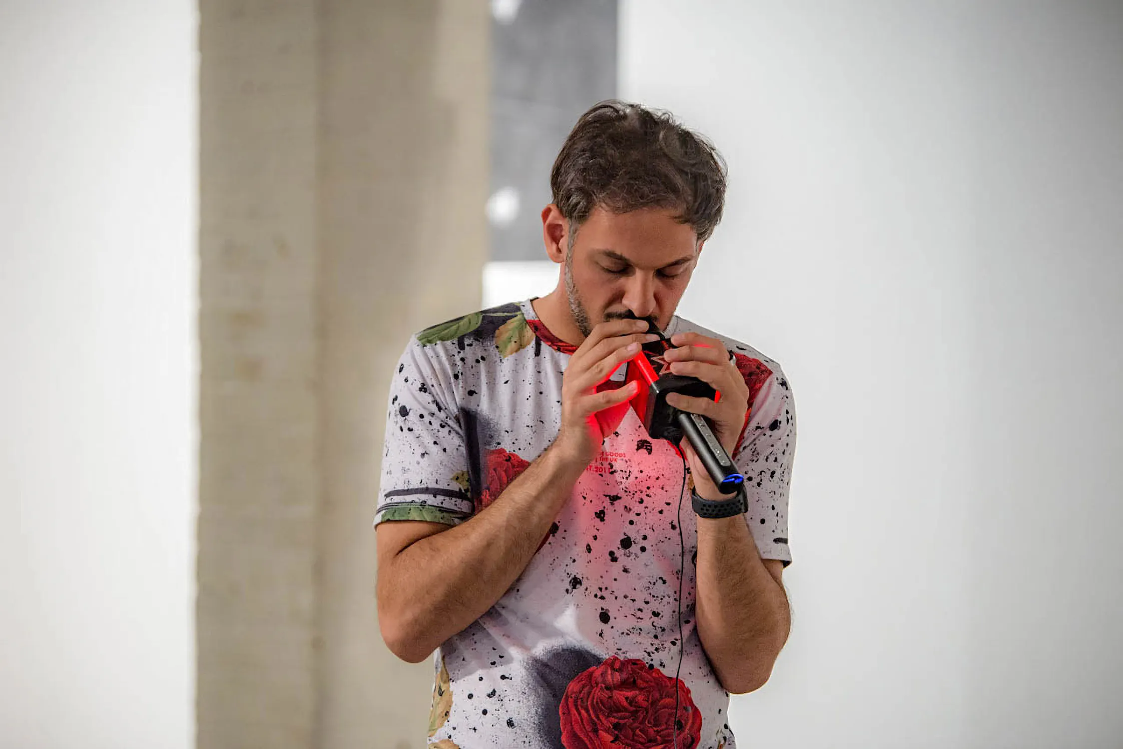 Abbas Zahedi, a man with short brown hair and a beard plays a small wind instrument into a microphone. He wears a white T-shirt with colourful paint splatters and a red rose print. The background is blurred, with white and beige walls.
