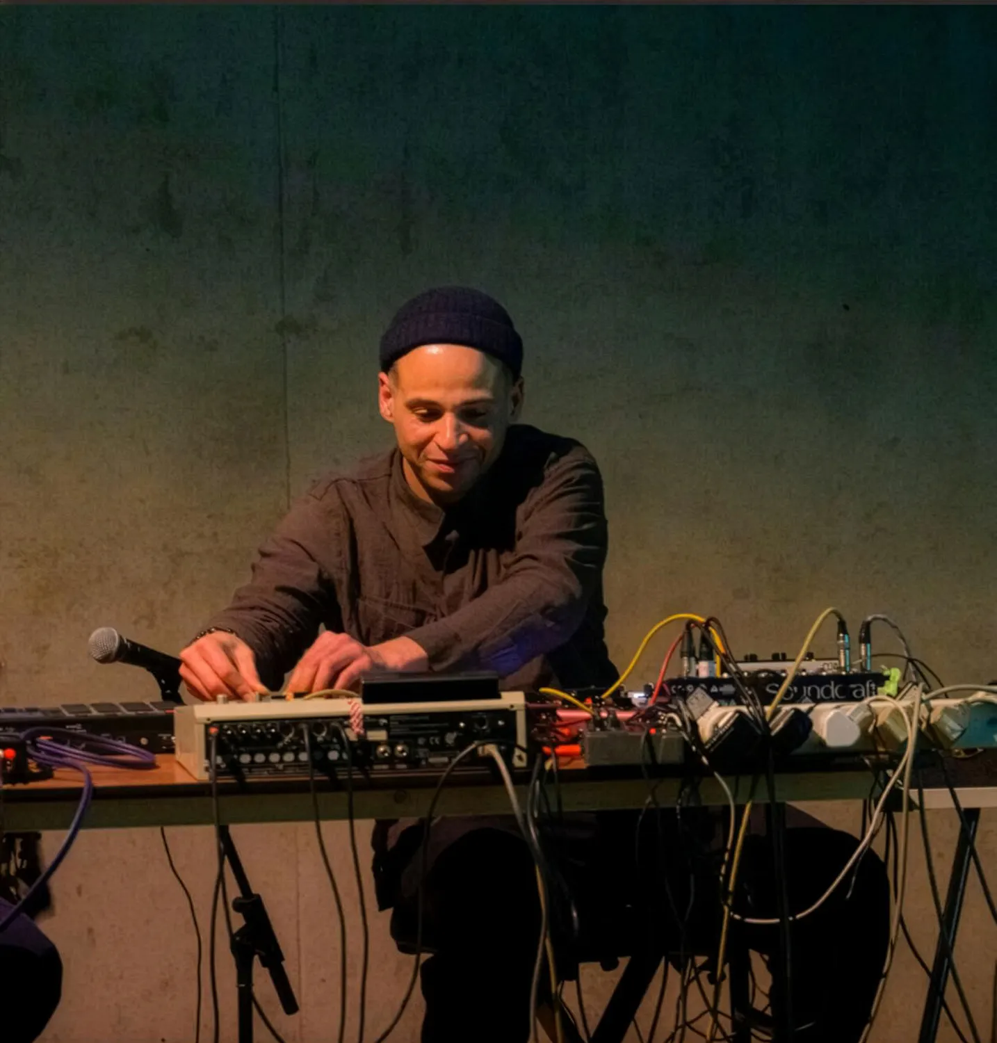 Tom Harris, a man wearing a dark beanie and jacket sits at a table with electronic music equipment and cables, adjusting controls. A microphone sits nearby. The background is plain and dimly lit, creating a focused, intimate atmosphere.