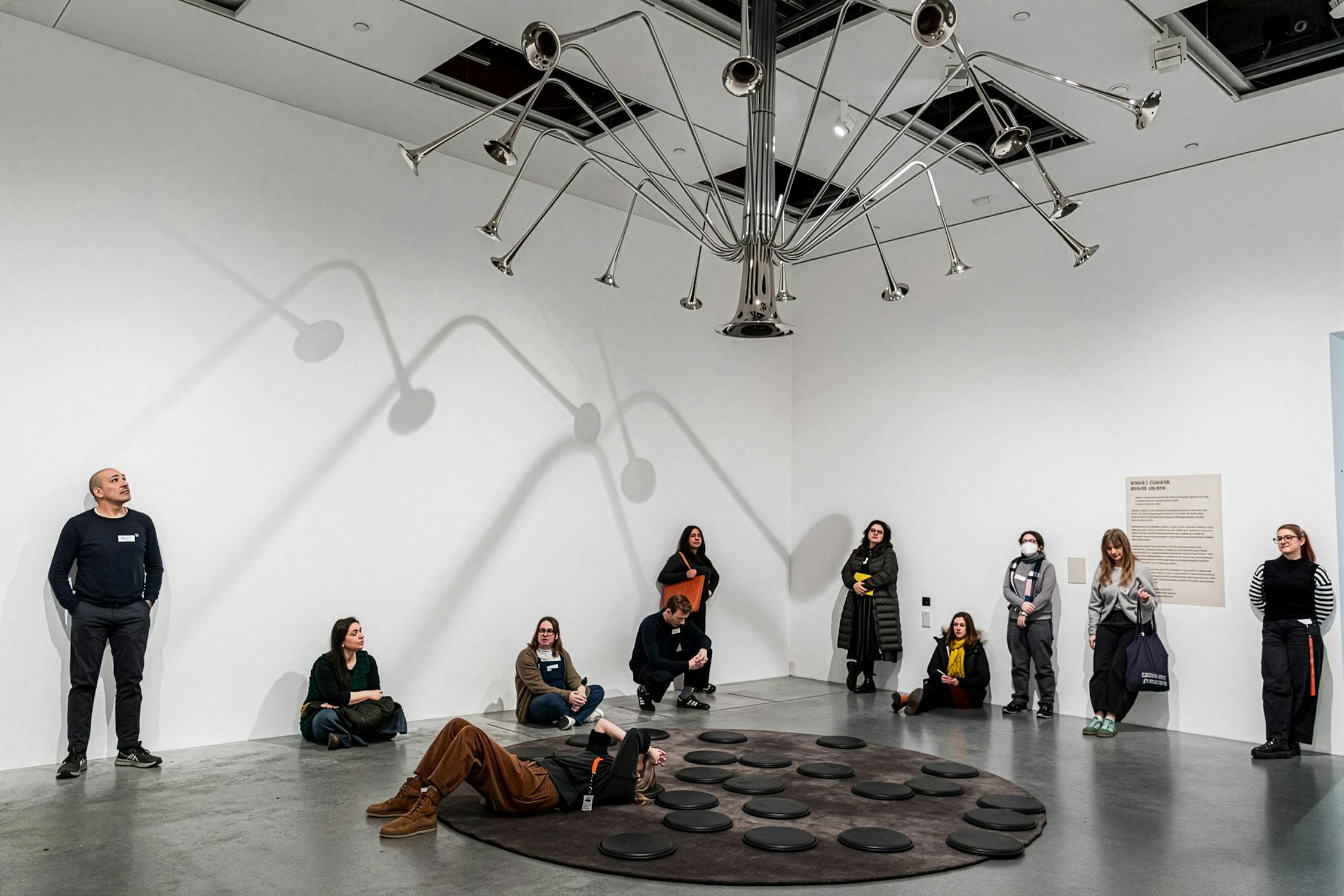 A group of people sit and stand in a white-walled gallery, observing a ceiling-mounted sculpture made of metal trumpets radiating outward. Shadows from the sculpture create patterns on the wall. Some people sit on black cushions; others stand nearby, facing the artwork.