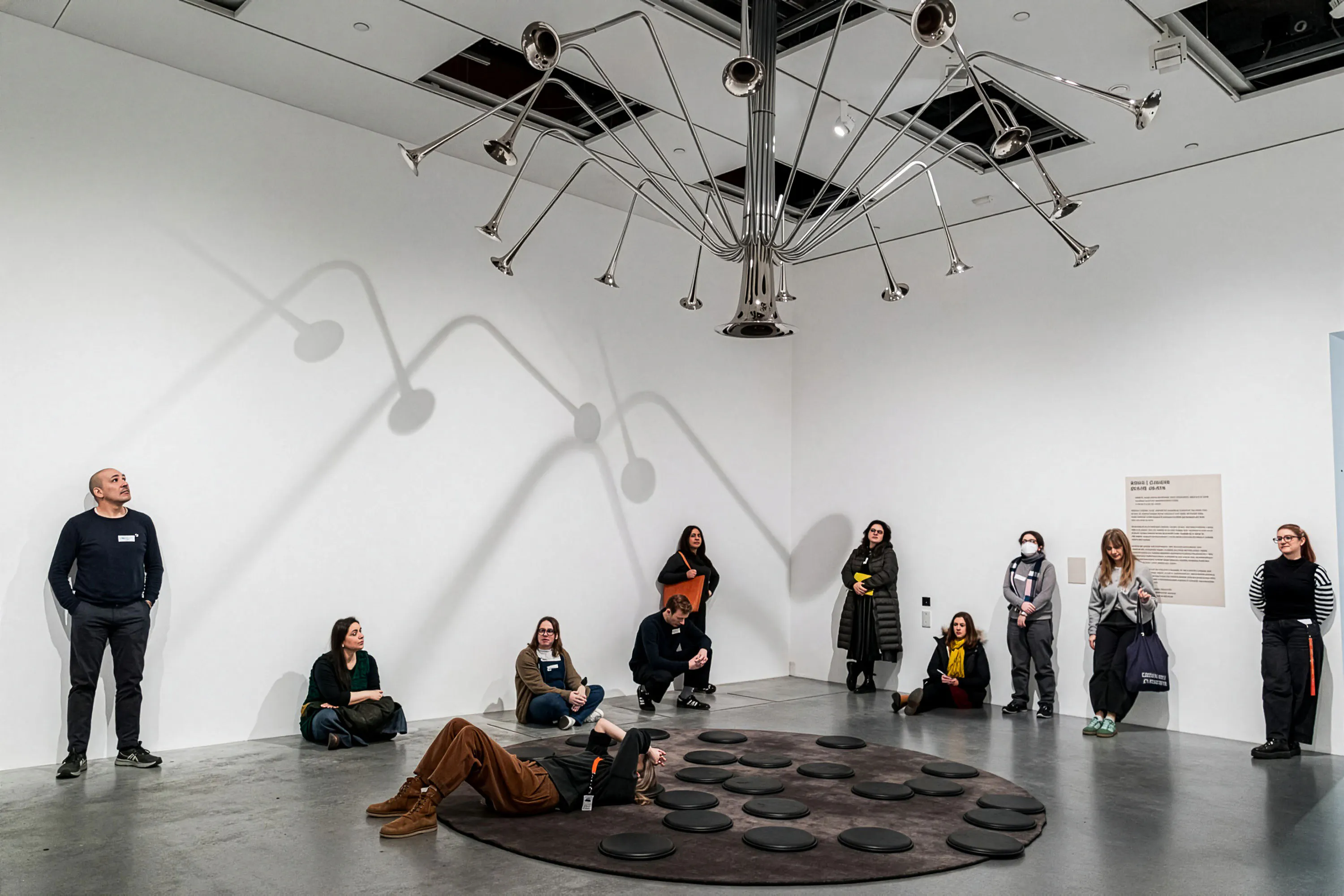 A group of people sit and stand in a white-walled gallery, observing a ceiling-mounted sculpture made of metal trumpets radiating outward. Shadows from the sculpture create patterns on the wall. Some people sit on black cushions; others stand nearby, facing the artwork.