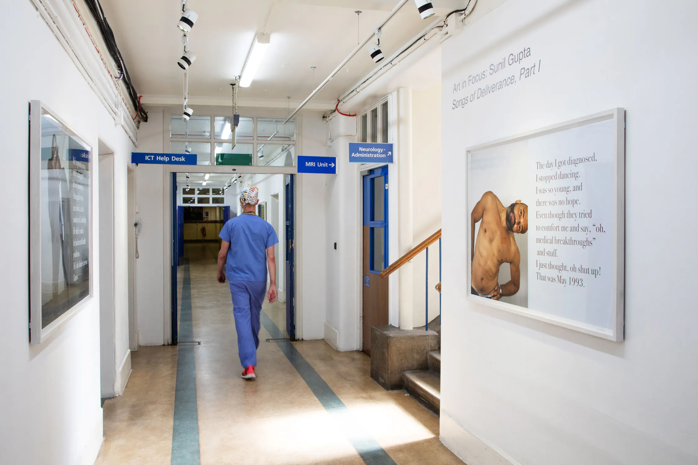 A person in blue scrubs and a surgical cap walks down a well-lit hospital corridor. Wall signs direct to various departments. Artworks, including a photo and text panel, hang on the white walls. The floor is beige, and there are overhead lights and pipes.