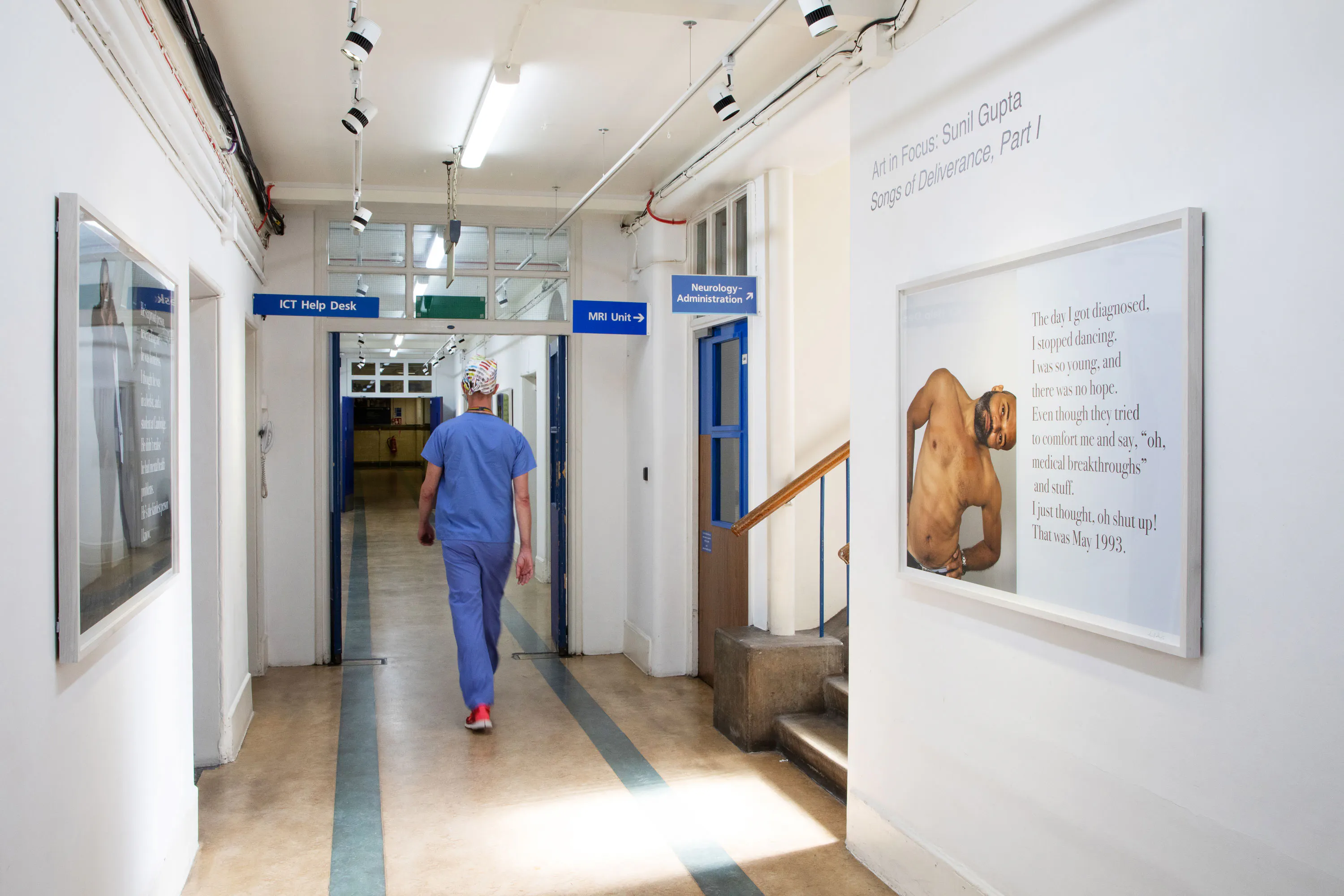 A person in blue scrubs and a surgical cap walks down a well-lit hospital corridor. Wall signs direct to various departments. Artworks, including a photo and text panel, hang on the white walls. The floor is beige, and there are overhead lights and pipes.