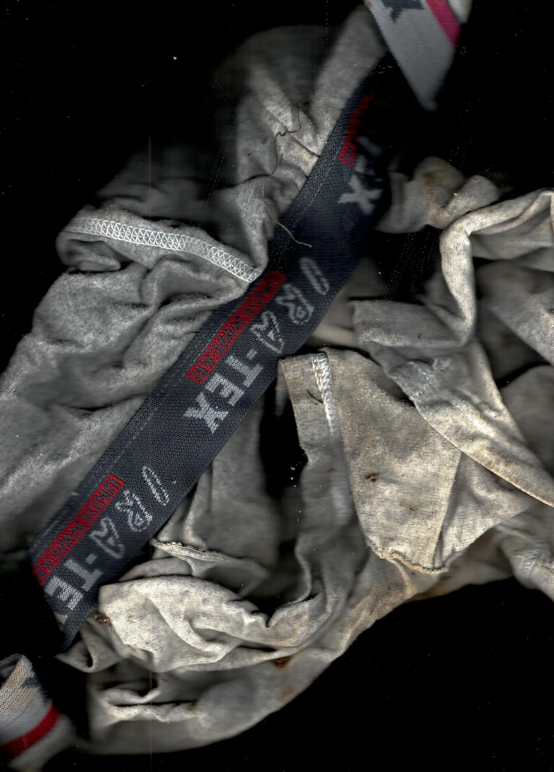 A close-up of crumpled, dirty grey underwear, partially revealing a dark blue elastic waistband with red and white text and symbols, including “4-TA-TI” and stars. The fabric appears stained and worn, set against a black background.