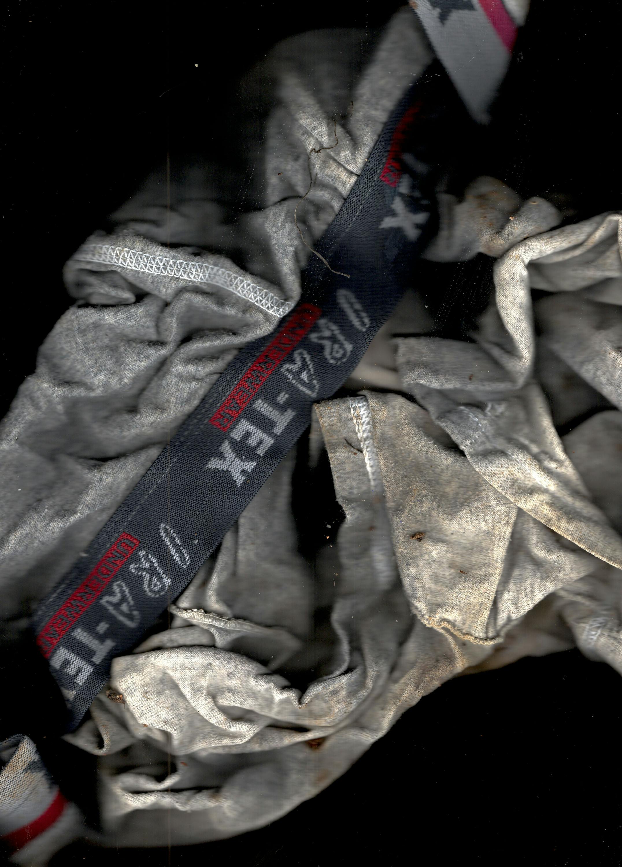 A close-up of crumpled, dirty grey underwear, partially revealing a dark blue elastic waistband with red and white text and symbols, including “4-TA-TI” and stars. The fabric appears stained and worn, set against a black background.