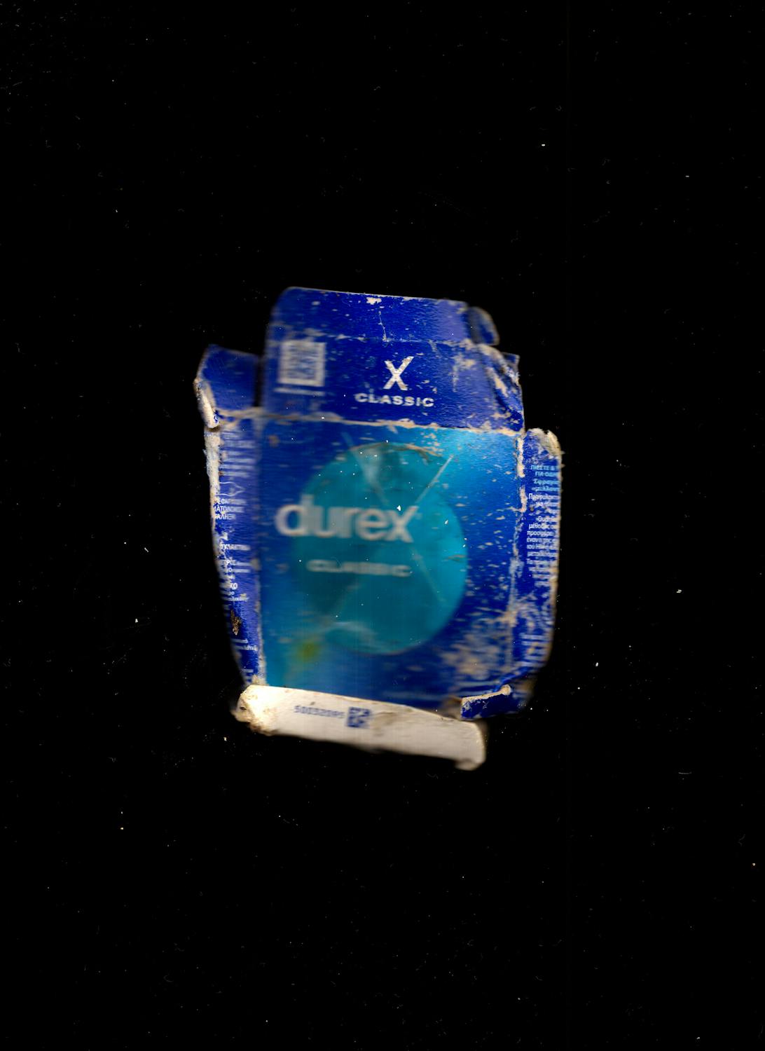 A battered, blue Durex Classic condom box is shown against a black background. The box is crumpled and worn, with visible creases, dirt, and faded areas, suggesting it has been discarded or exposed to the elements.