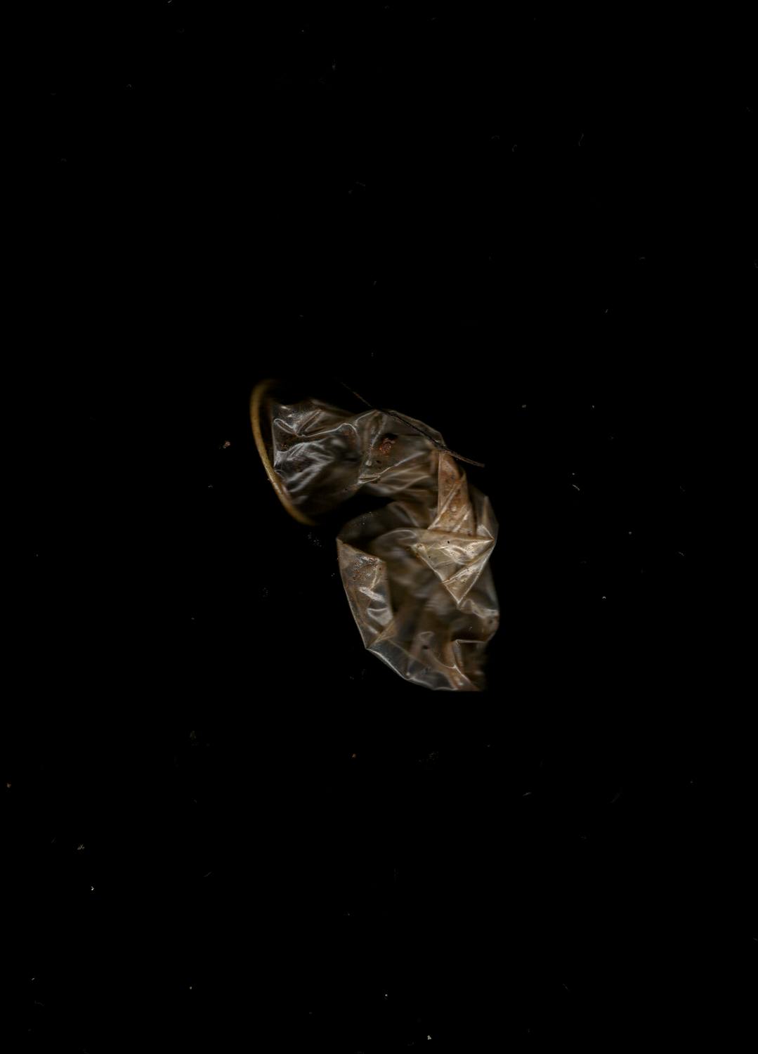 A single, crumpled, transparent condom is centred on a solid black background. The condom is folded and creased. The lighting emphasises the object's shiny, reflective surface while the background remains completely dark.