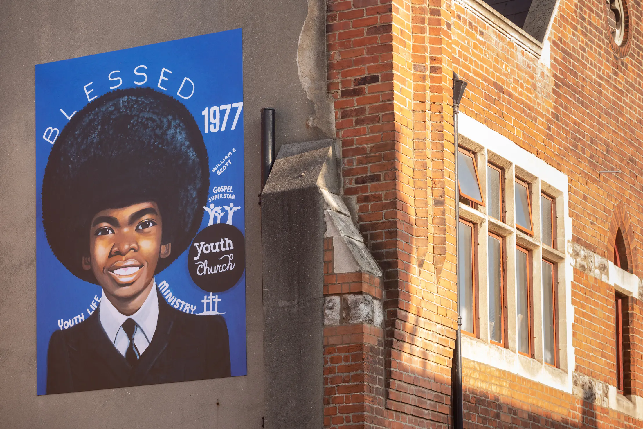 A William Scott mural on a brick building shows a smiling Black boy with a large afro, wearing a suit and tie. The mural reads “BLESSED 1977,” “Youth Life Ministry,” and other text. Sunlit bricks and windows of the neighbouring building are also visible.