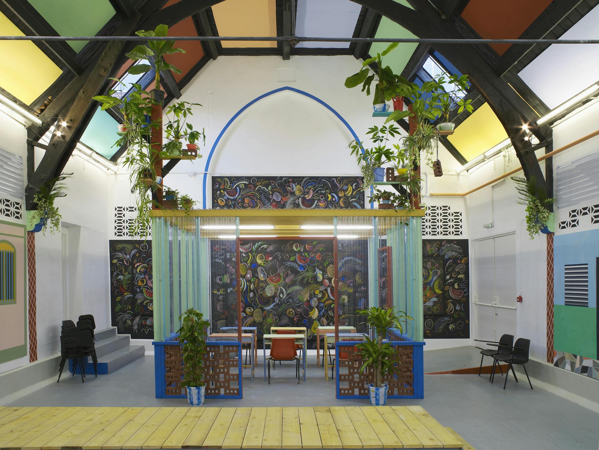 A view of Sol Calero’s immersive installation at Studio Voltaire, presenting a bright, modern room with high arched ceilings, hanging plants, glass-panelled partitions, and a central table surrounded by mismatched chairs. The walls are adorned with intricate artwork and colourful panels, contributing to the space’s playful and collaborative feel.