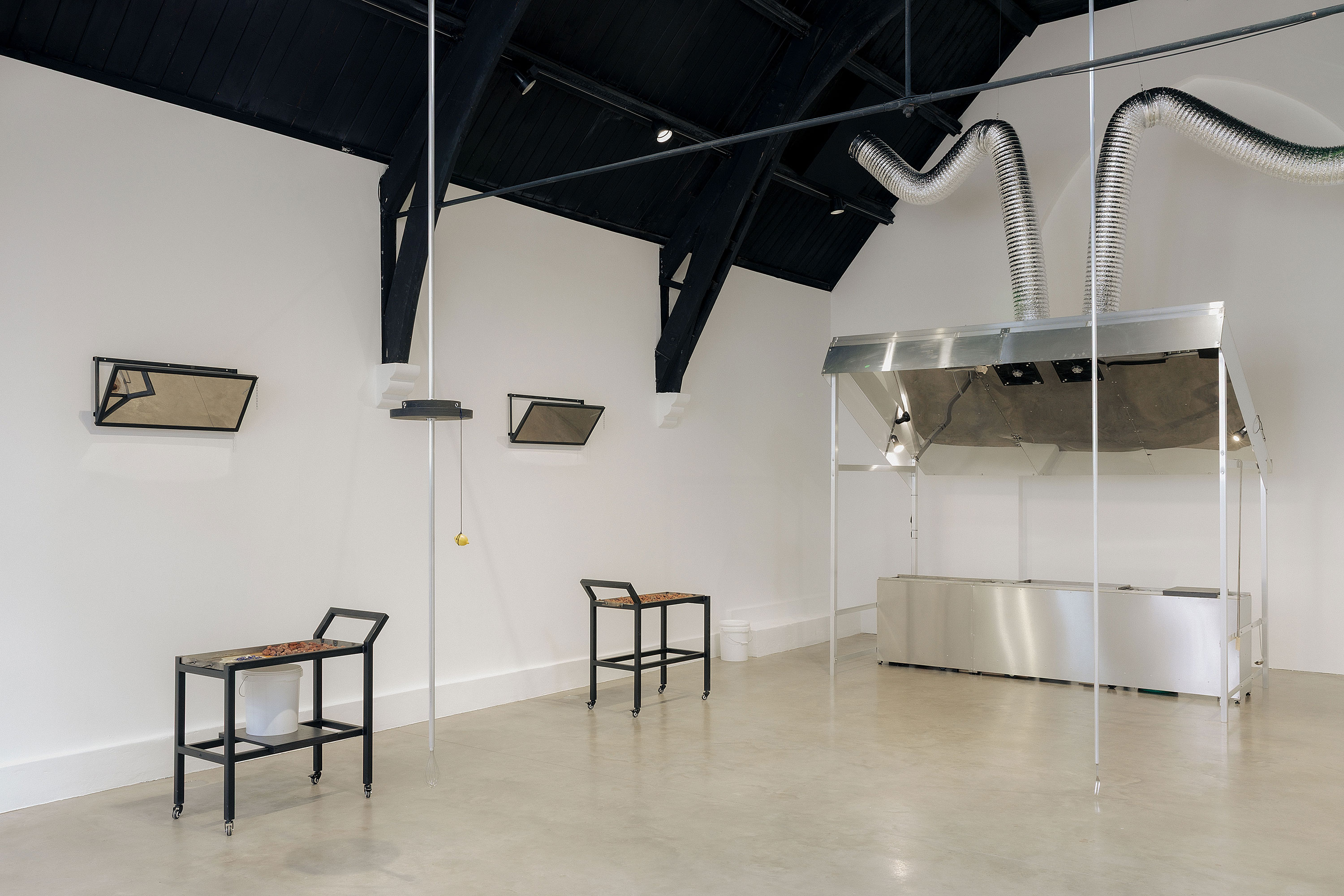 A contemporary gallery space with high black ceilings, white walls, and polished concrete floors. Two small tables and chairs face each other; mirrored boxes are mounted on the walls, and a metal hood with flexible ducts hangs in the background.