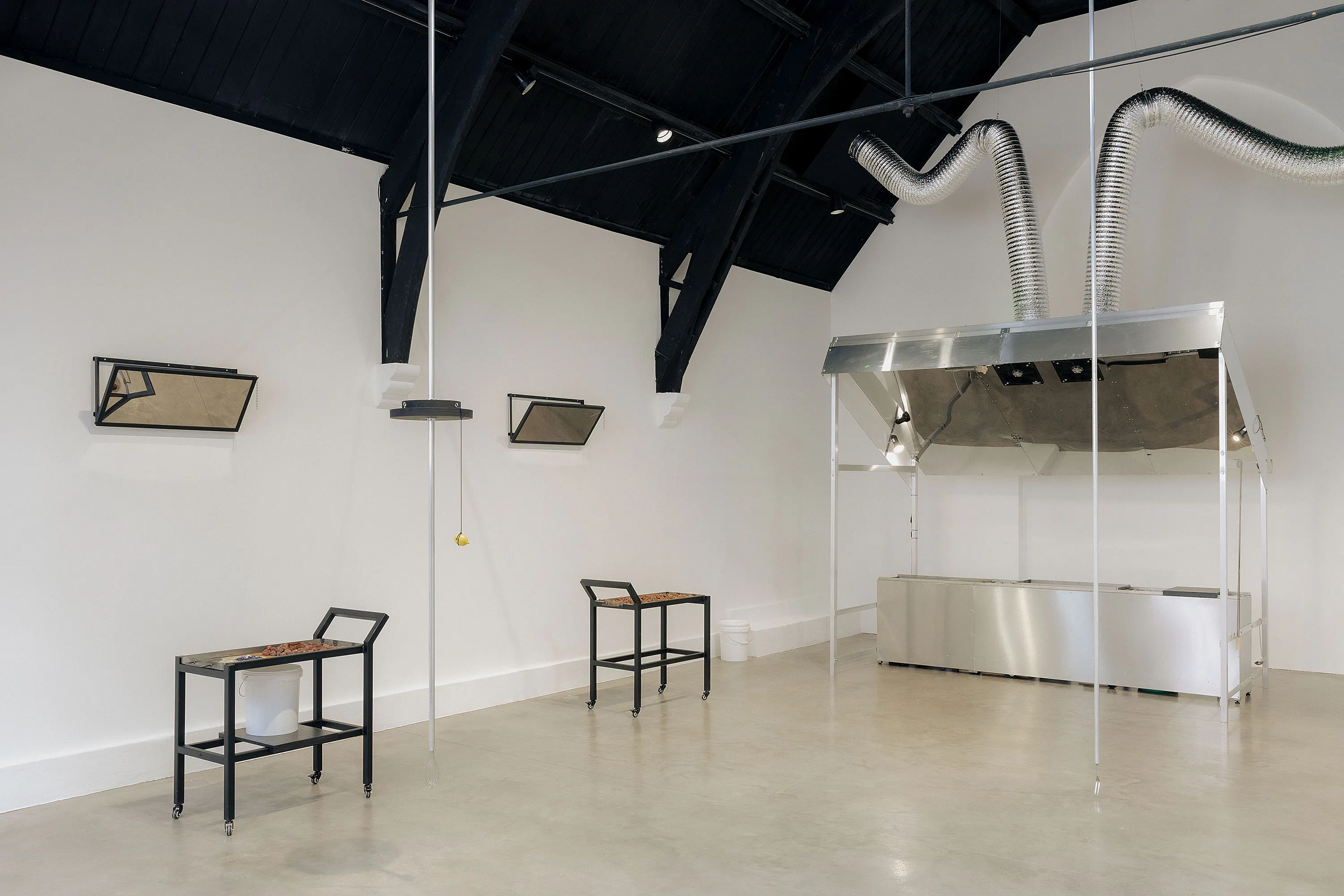 A contemporary gallery space with high black ceilings, white walls, and polished concrete floors. Two small tables and chairs face each other; mirrored boxes are mounted on the walls, and a metal hood with flexible ducts hangs in the background.