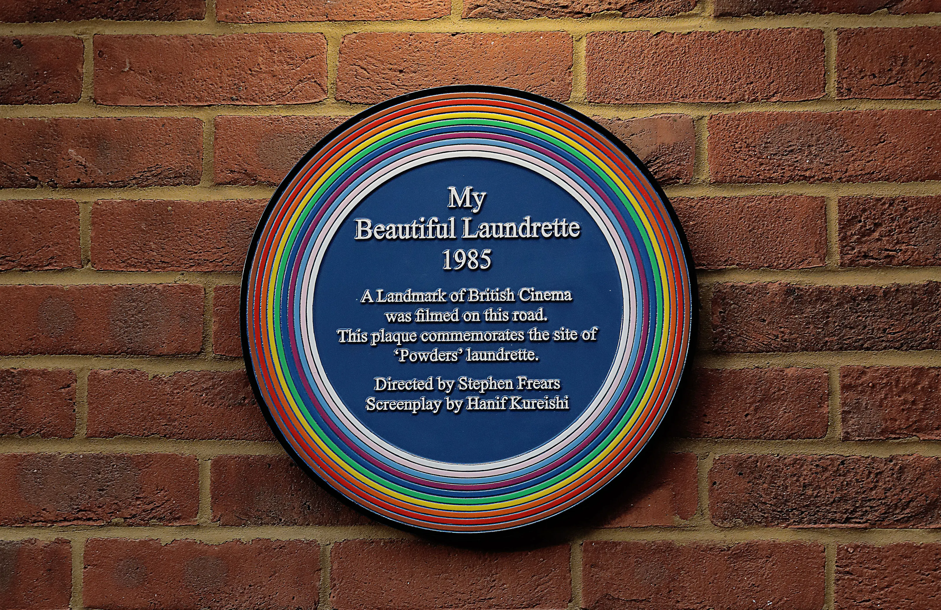 A circular blue plaque with a multicoloured ring border is mounted on a brick wall. The plaque commemorates the filming of "My Beautiful Laundrette" (1985) and credits Stephen Frears (director) and Hanif Kureishi (screenplay).