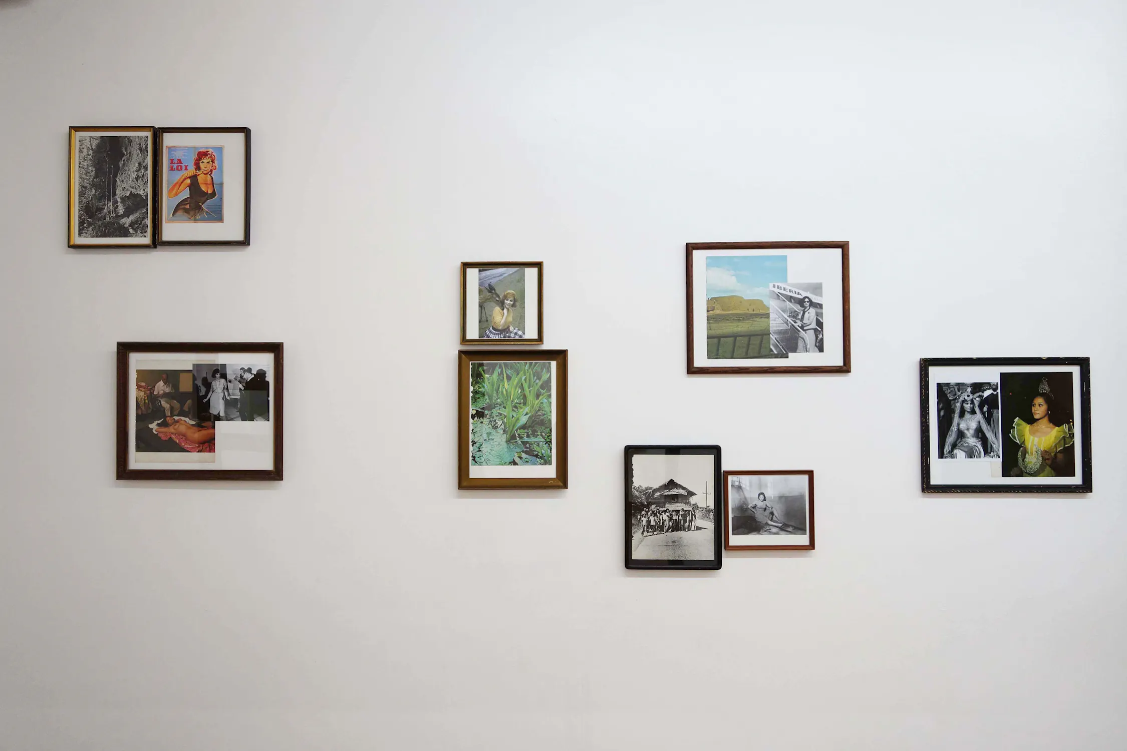 A white wall displays eight framed photos and artworks by Mark Salvatus, in varying sizes and colours, arranged in an irregular grid. The images include portraits, landscapes, and nature scenes, each with a unique frame, creating an eclectic, artistic gallery display.
