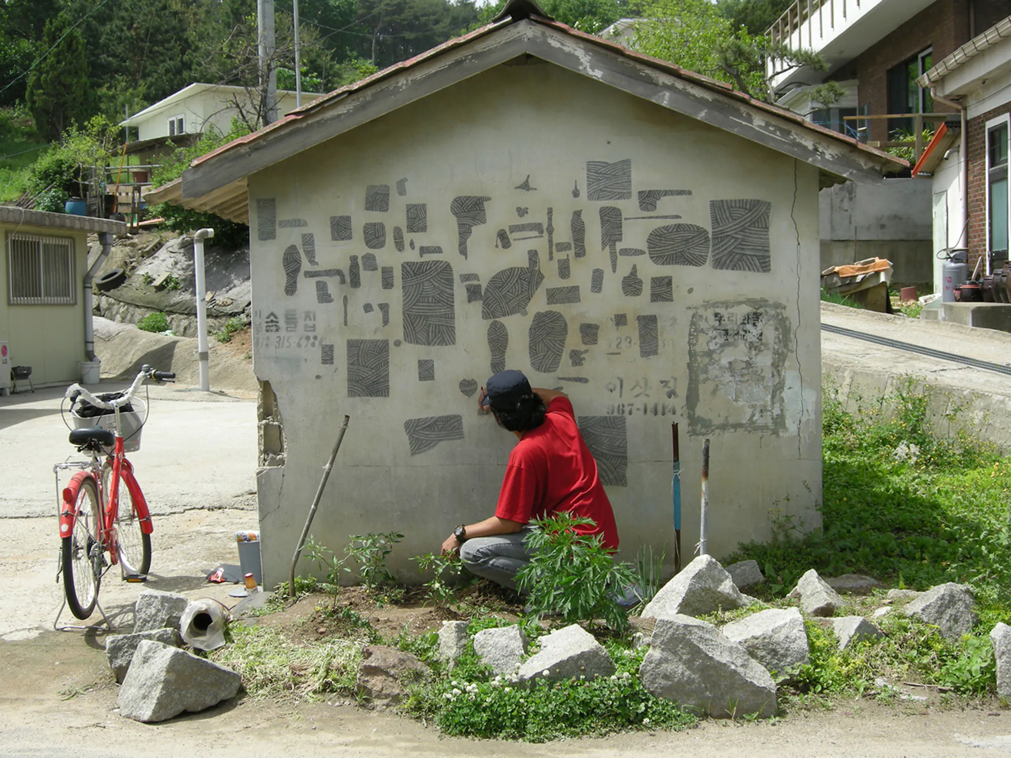 A person in a red shirt squats by a small building, drawing abstract shapes and patterns on its wall. A red bicycle is parked nearby, and scattered rocks and greenery surround the area. The setting appears residential and peaceful.