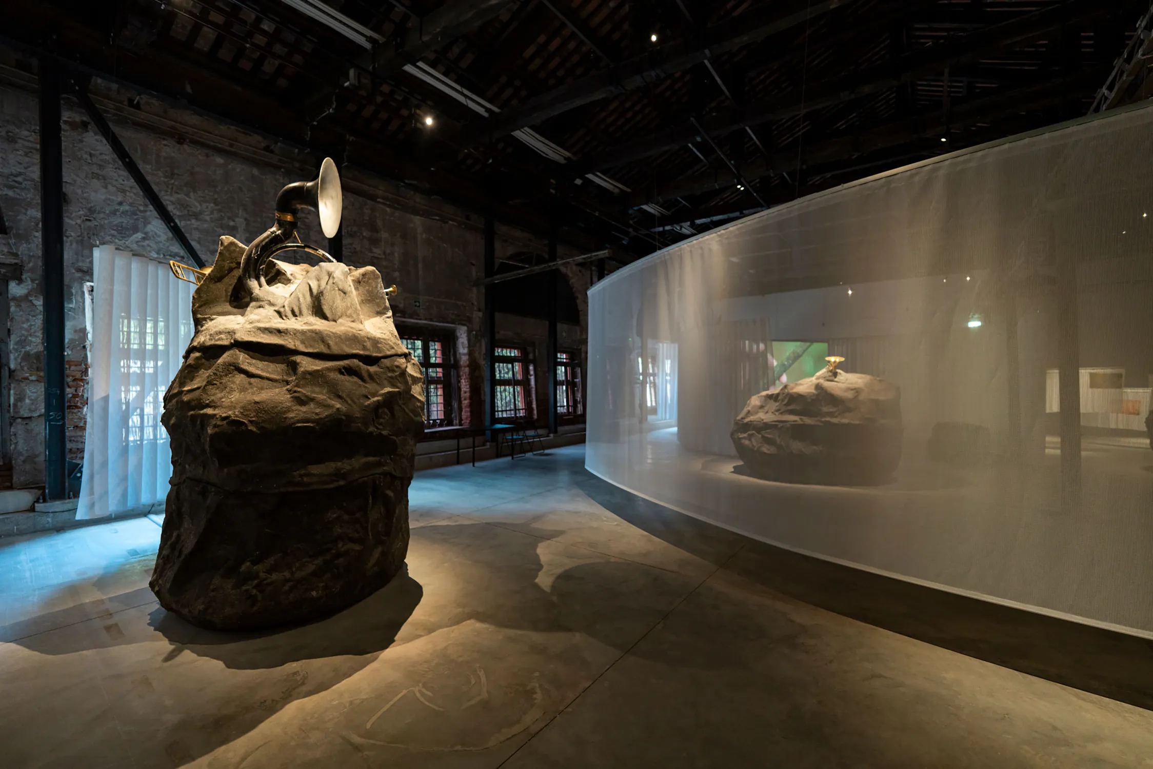 A dimly lit art gallery features two large rock-like sculptures by Mark Salvatus. One in the foreground has a brass horn and silver dish attached. The other sits behind sheer white fabric. Exposed beams and brick walls surround the spacious, modern industrial setting.