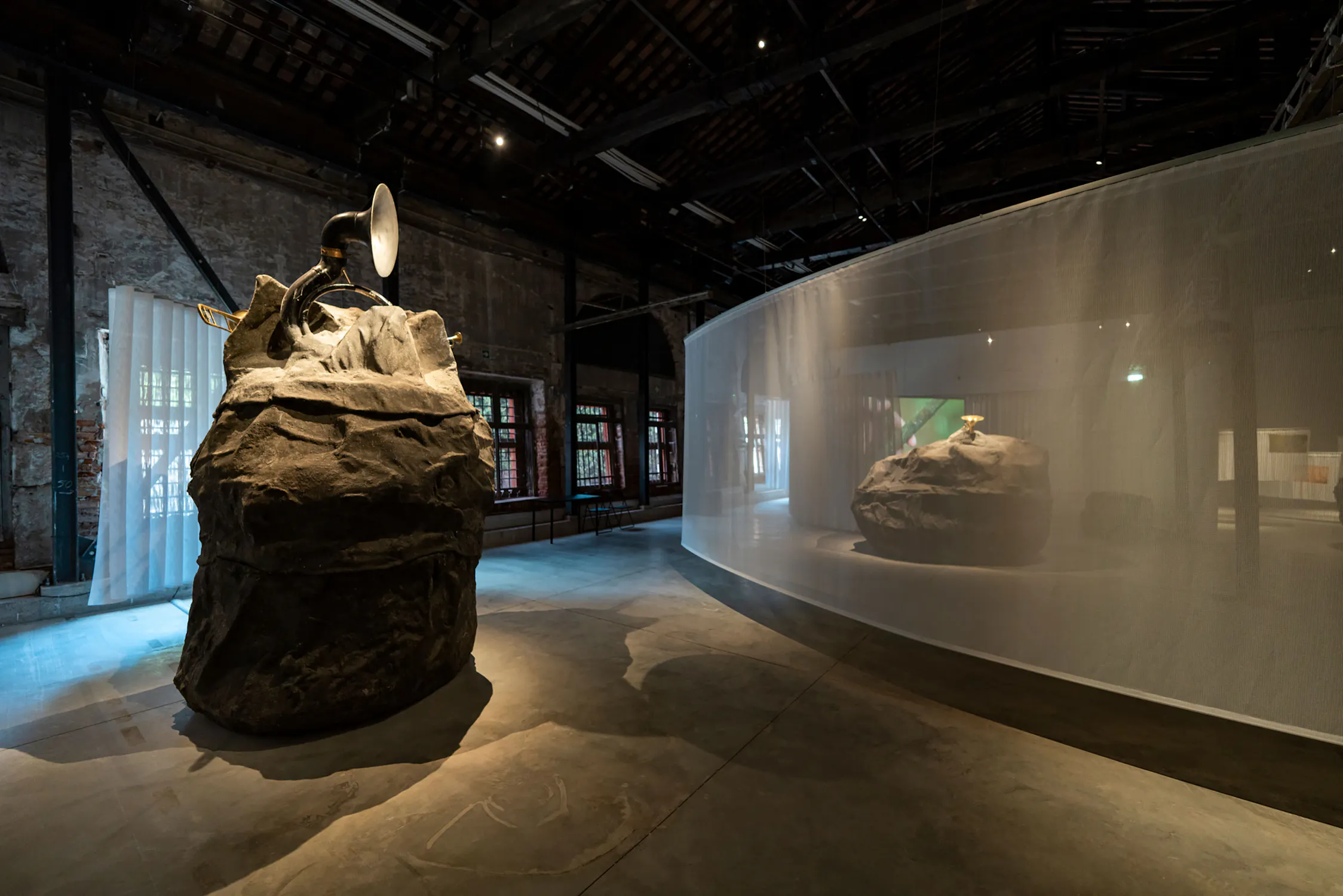 A dimly lit art gallery features two large rock-like sculptures by Mark Salvatus. One in the foreground has a brass horn and silver dish attached. The other sits behind sheer white fabric. Exposed beams and brick walls surround the spacious, modern industrial setting.