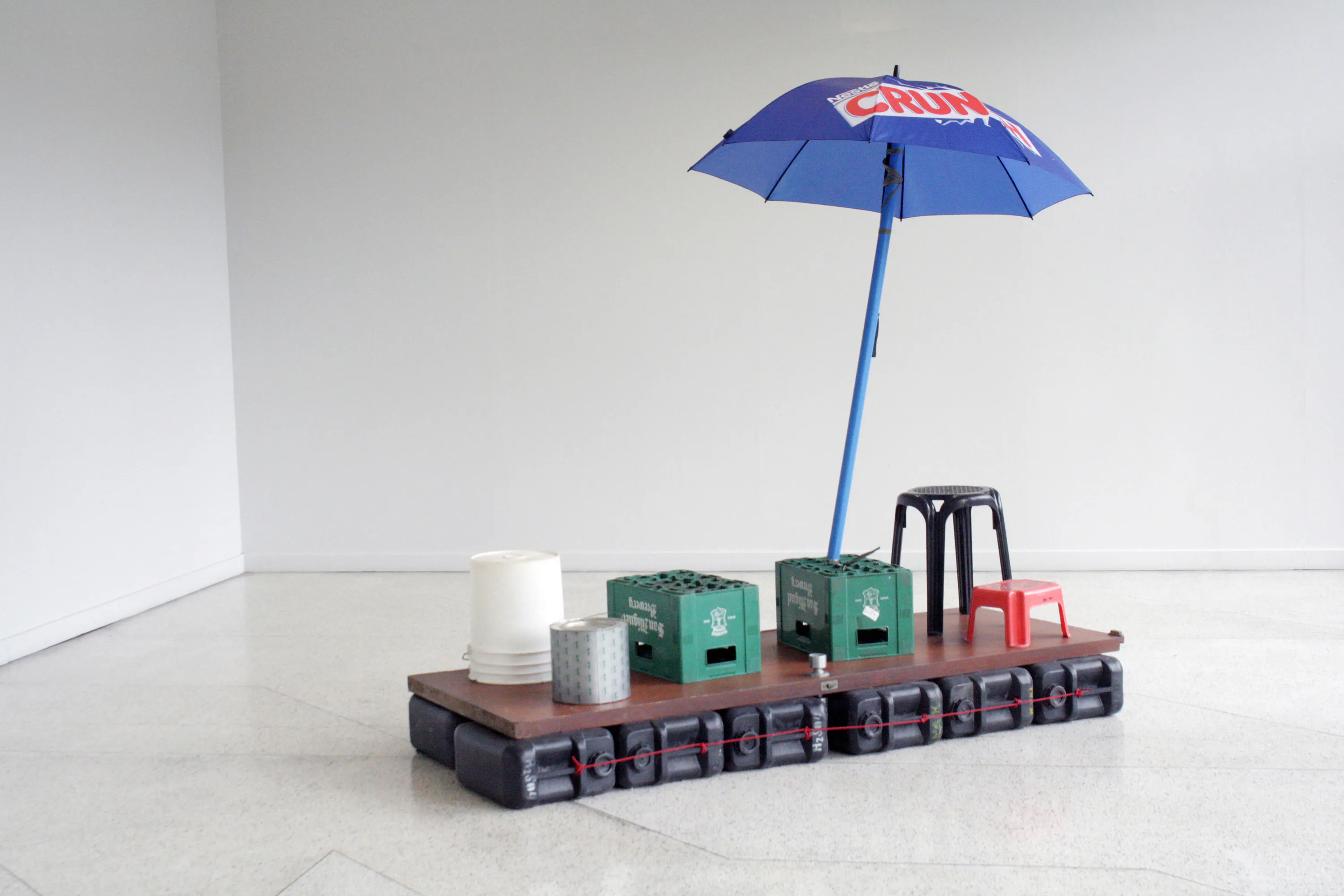A floating platform made of black containers supports a wooden surface with two green crates, a white bucket, a metal tin, black and red plastic stools, and a large blue umbrella with "CRUX" text, set in a bare, white room.