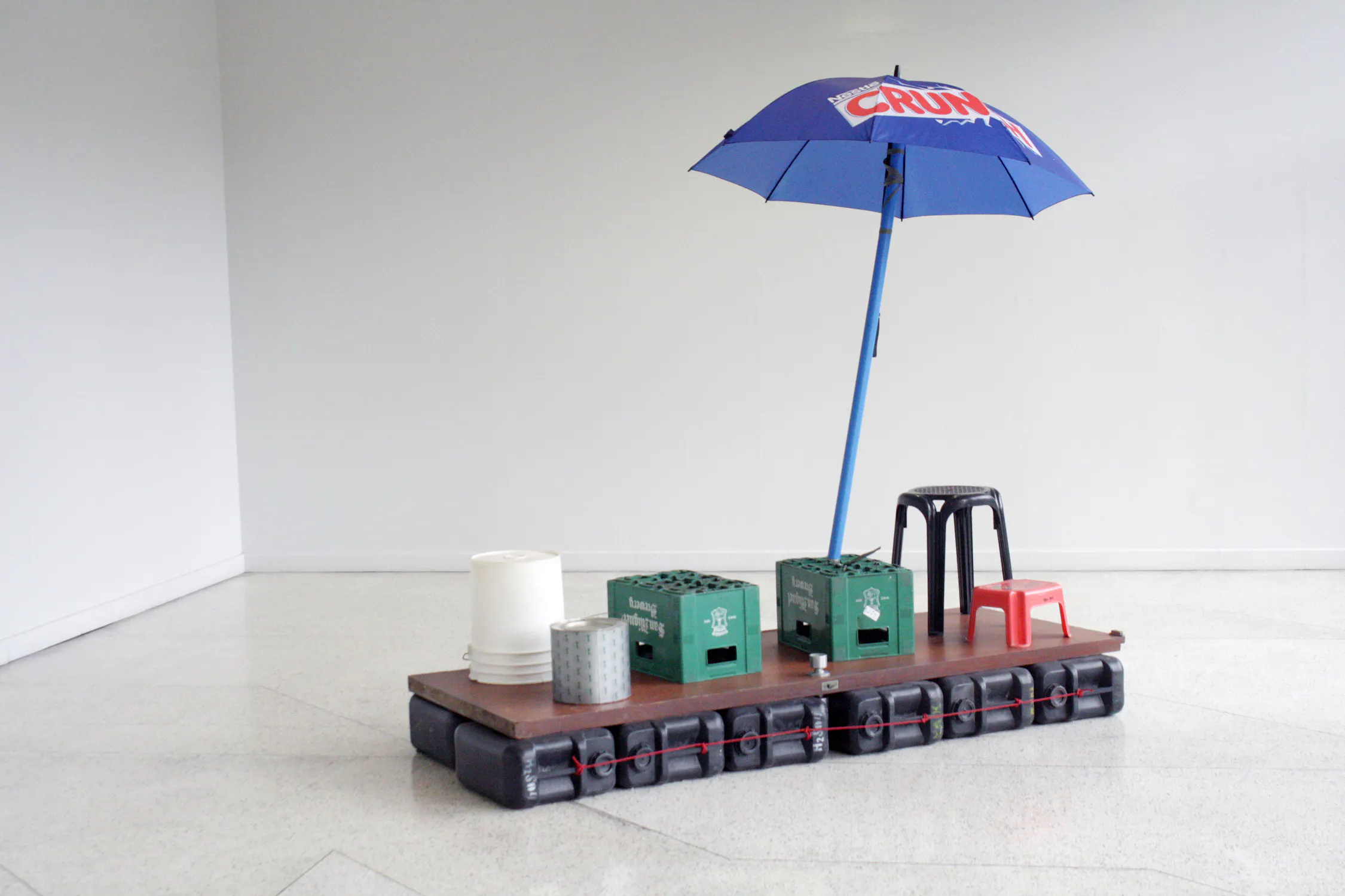 A floating platform made of black containers supports a wooden surface with two green crates, a white bucket, a metal tin, black and red plastic stools, and a large blue umbrella with "CRUX" text, set in a bare, white room.