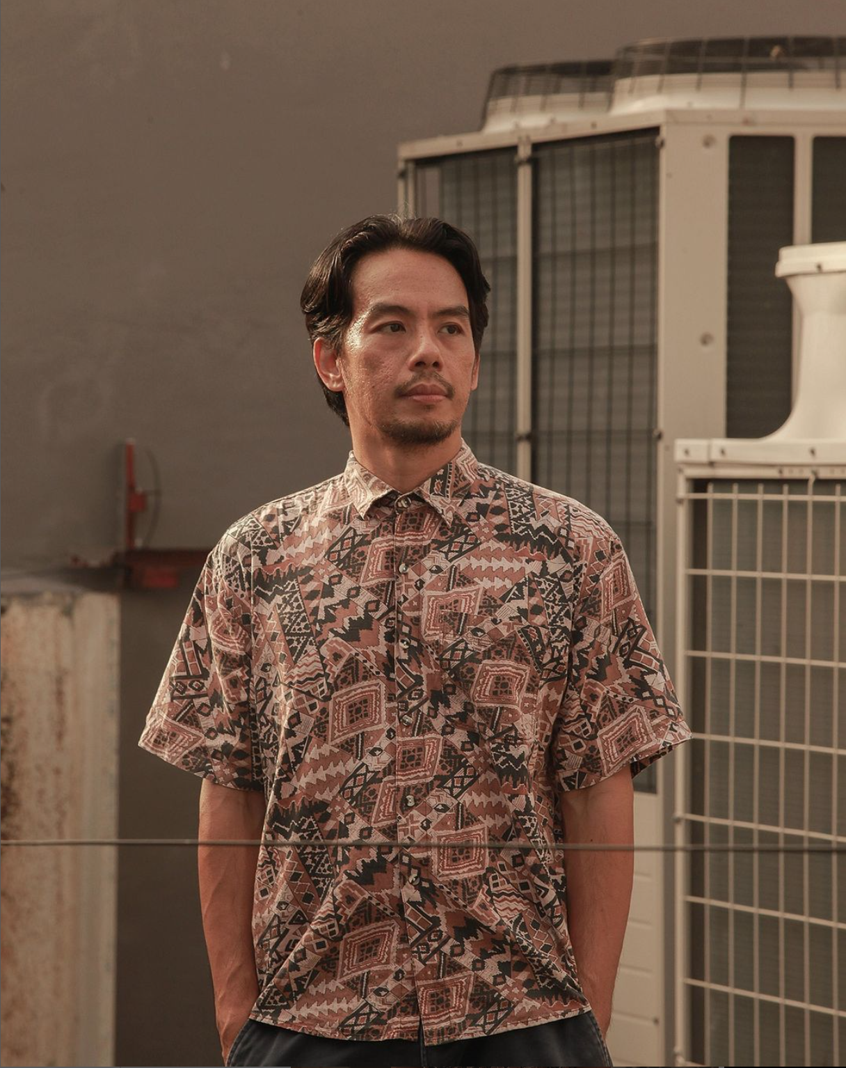 Artist Mark Salvatus: a man with short dark hair and a trimmed beard wears a patterned short-sleeve shirt, standing with his hands in his pockets. He looks to the side. Behind him are large industrial air-conditioning units and a concrete wall, lit neutrally.