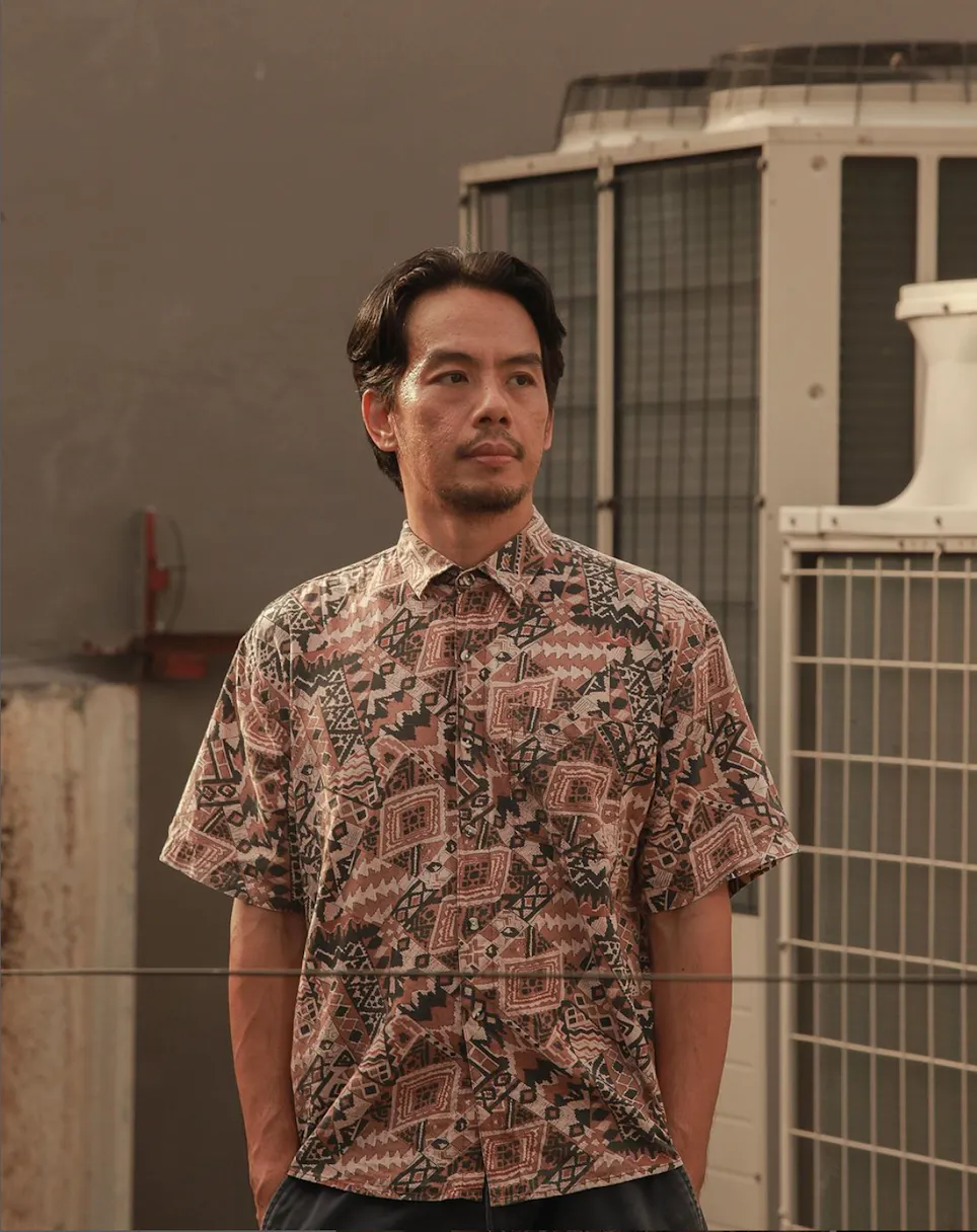 Artist Mark Salvatus: a man with short dark hair and a trimmed beard wears a patterned short-sleeve shirt, standing with his hands in his pockets. He looks to the side. Behind him are large industrial air-conditioning units and a concrete wall, lit neutrally.