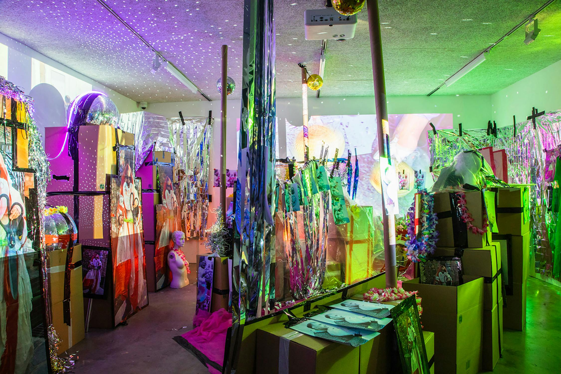 A vibrant art installation featuring an array of colorful lights, reflective materials, and creative decorations. Cardboard boxes are stacked alongside various objects under multicoloured lighting, creating an immersive, eclectic atmosphere.