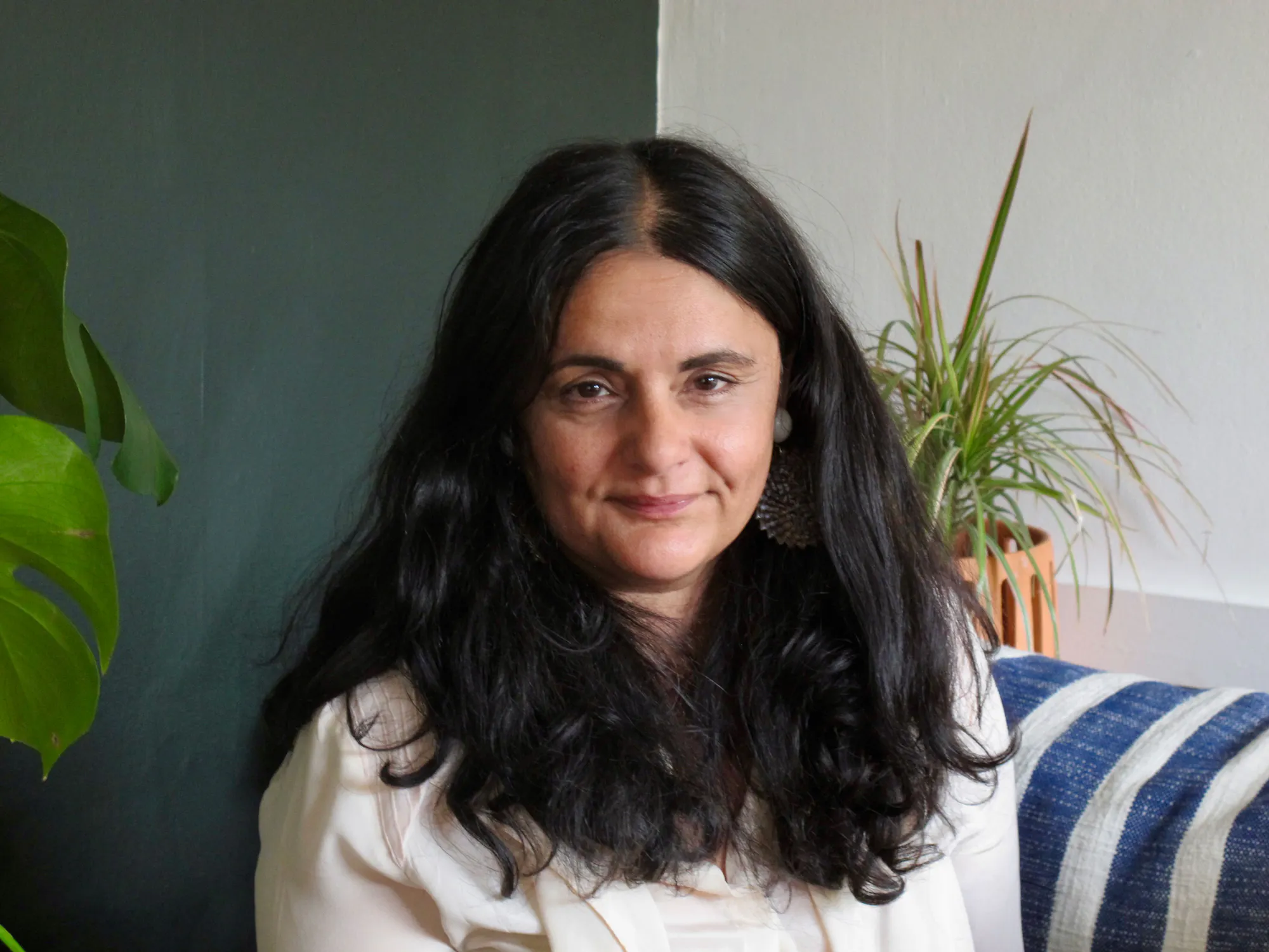 Habda Rashid is seated indoors, smiling softly. She wears a white blouse and large earrings. Behind her are green plants and a white-and-blue striped cushion. The wall is two-toned, dark green on the left and white on the right.