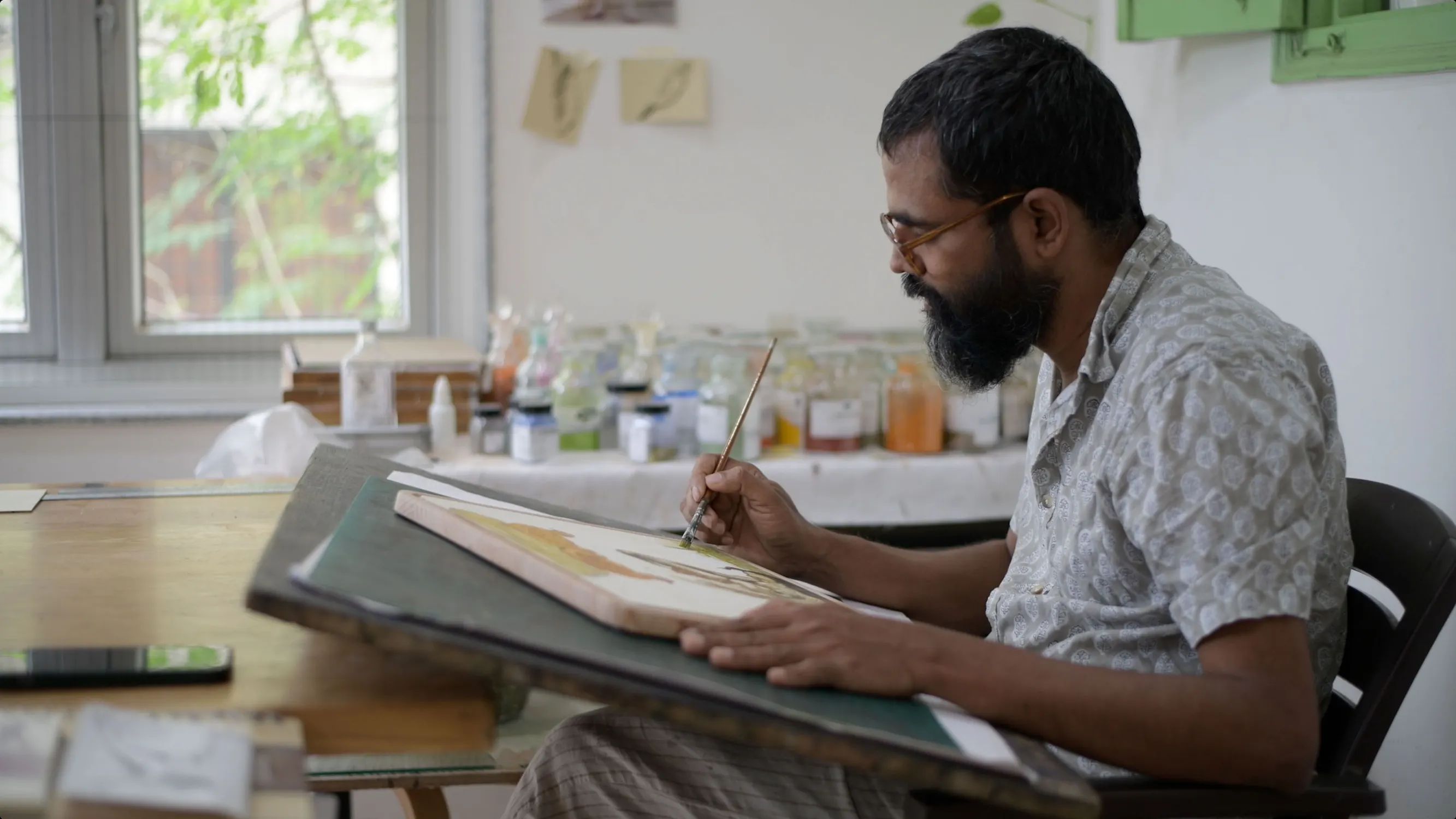 T Venkanna sits at a desk, carefully painting on a large sheet of paper on a tilted drawing board. Art supplies and jars are arranged on a table by a bright window in the background, creating a calm, creative workspace.