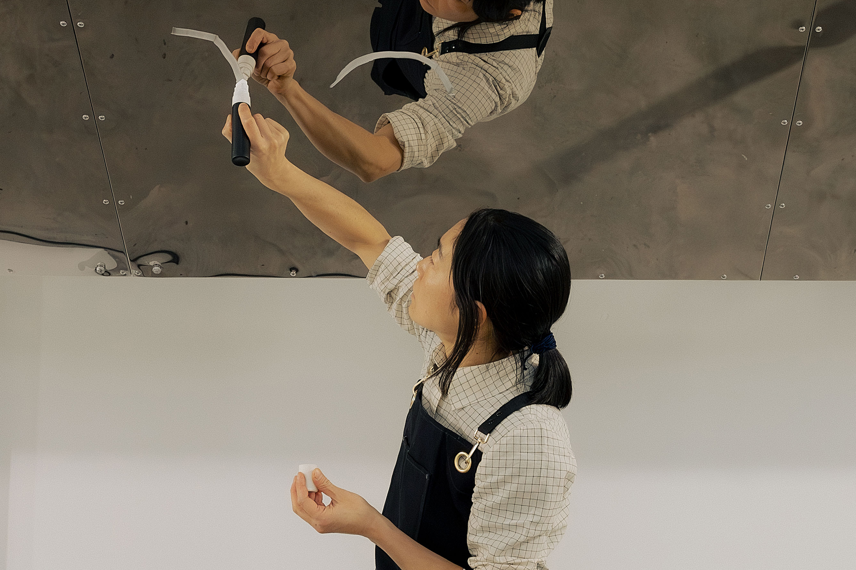 Aki Sasamoto wearing a checked shirt and dark dungarees, applies white pen to a shiny metal surface above them. Their reflection is visible on the metal. They hold more tape in their left hand and look upward, focusing on their task.