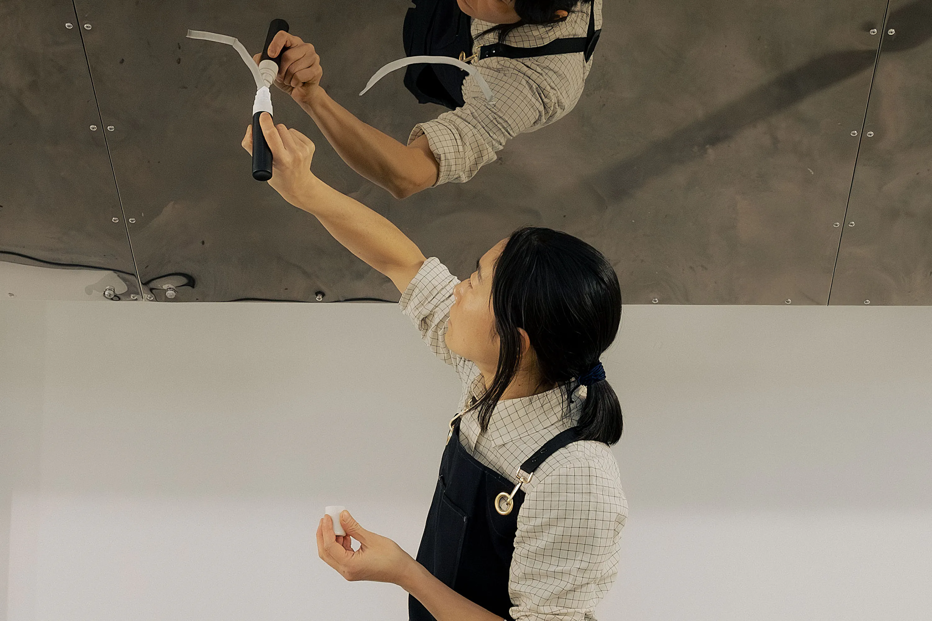 Aki Sasamoto wearing a checked shirt and dark dungarees, applies white pen to a shiny metal surface above them. Their reflection is visible on the metal. They hold more tape in their left hand and look upward, focusing on their task.