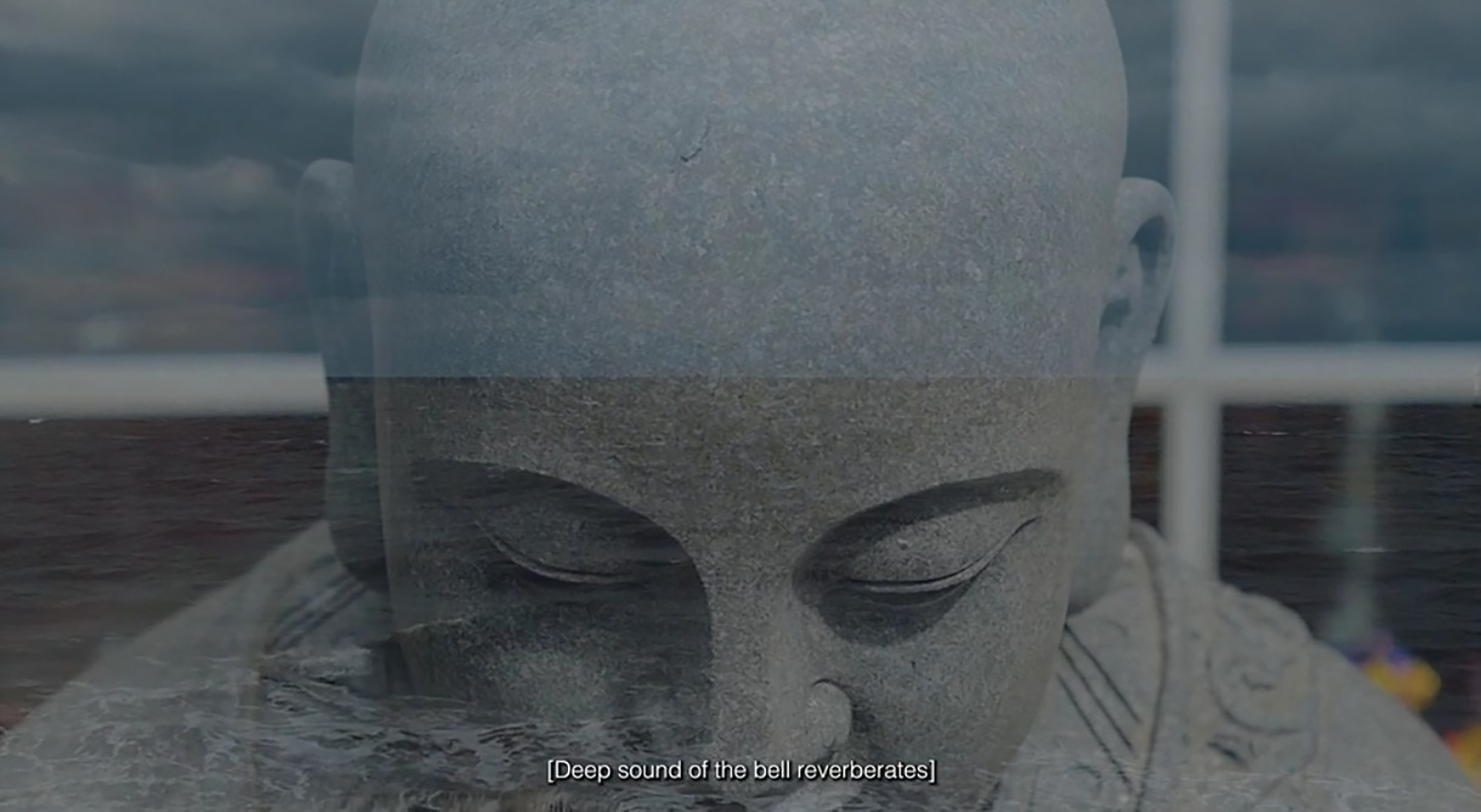 A close-up of a serene stone statue’s face, eyes closed, is overlaid with an image of calm, rippling ocean water. White subtitle text at the bottom reads: “[Deep sound of the bell reverberates]”. The mood is peaceful and contemplative.
