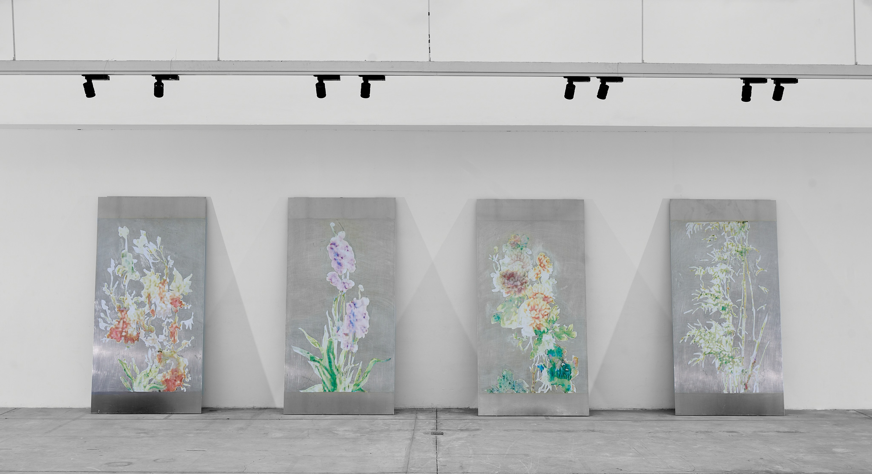 Three large, rectangular metal panels with delicate, pastel-coloured botanical illustrations stand upright against a white gallery wall under black track lights. The flowers and plants on each panel differ in shape and colour, with a concrete floor below.