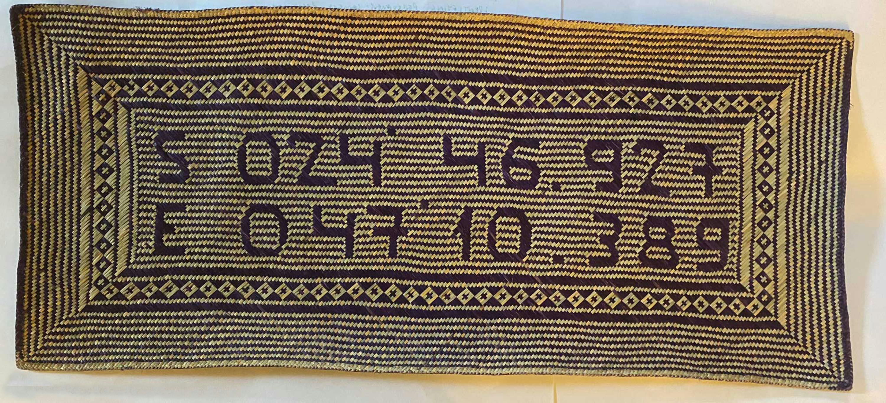 A rectangular woven mat with black and tan stripes, diamond patterns along the borders, and abstract geometric shapes and lines in the centre, possibly representing stylised numbers or symbols.
