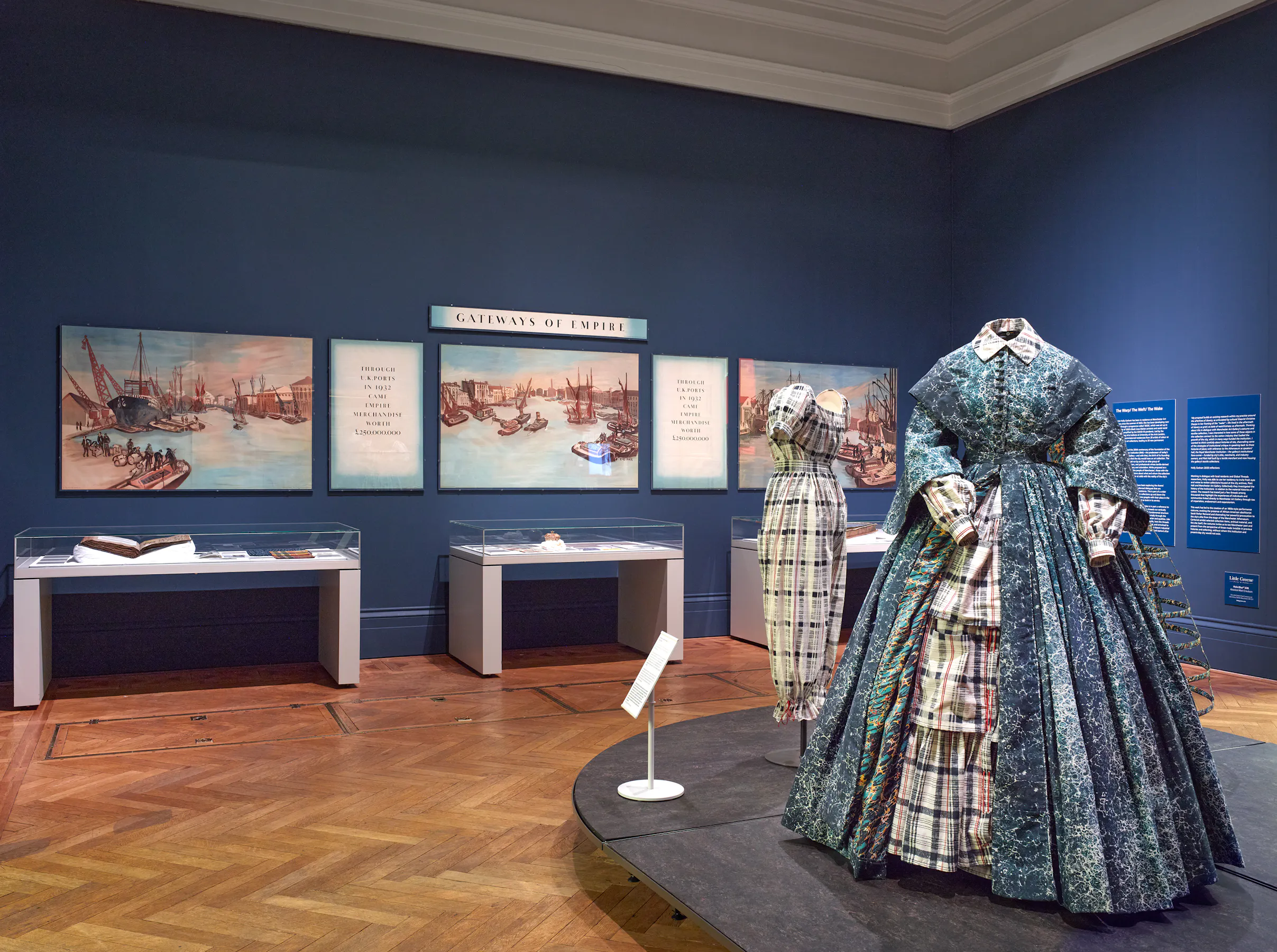 An exhibition by Holly Graham with blue walls displays historical dresses on mannequins, glass cases with books and artefacts, and three paintings of ships and harbours. A sign reads “Gateways of Empire” above the paintings, and an exhibition panel is mounted on the right wall.